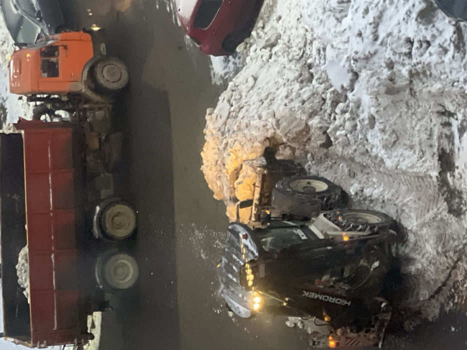 A nighttime scene captures construction vehicles amid snowy conditions. A large orange dump truck is visible. Partially submerged in water or slush, a backhoe with "HIDROMEK" on its side has its headlights illuminated, positioned near a pile of snow. The surroundings reflect the lights of the vehicles, indicating a wet surface. A red car is also parked near the area.
