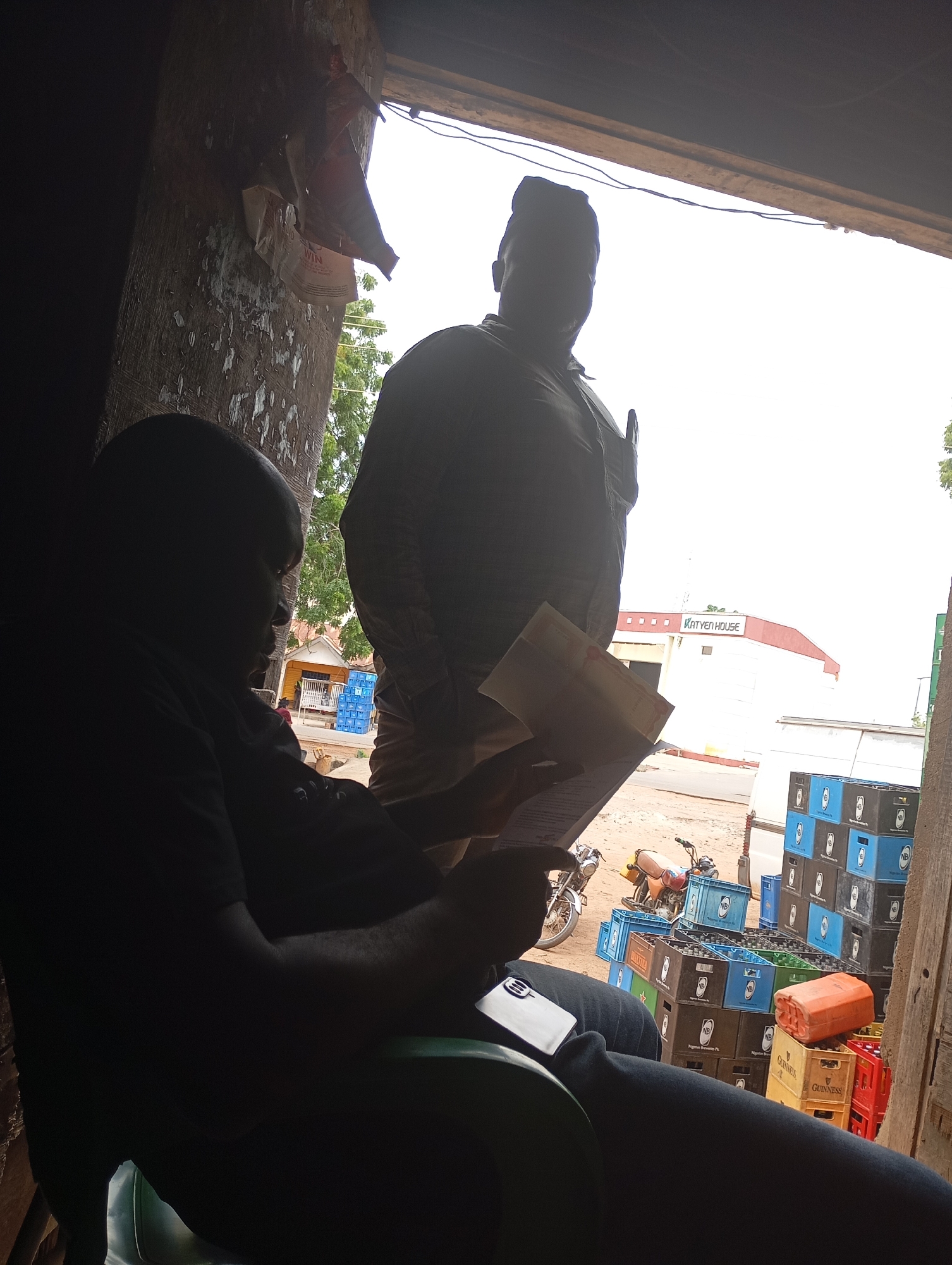 The image depicts a scene at an outdoor establishment in Shendam, Nigeria, likely a local bar or shop. In the foreground, a person is seated, silhouetted against the bright outdoor light, engrossed in reading a document. Another individual stands behind them, also silhouetted, wearing a cap and what appears to be a uniform shirt.
The environment is rustic, with wooden structures and peeling paint visible on a pillar. Outside, a building with a sign that reads "KATYEN HOUSE" is visible, along with stacks of colorful crates, some filled with bottles. A motorcycle is parked nearby. The overall impression is a casual, everyday setting in a Nigerian city.