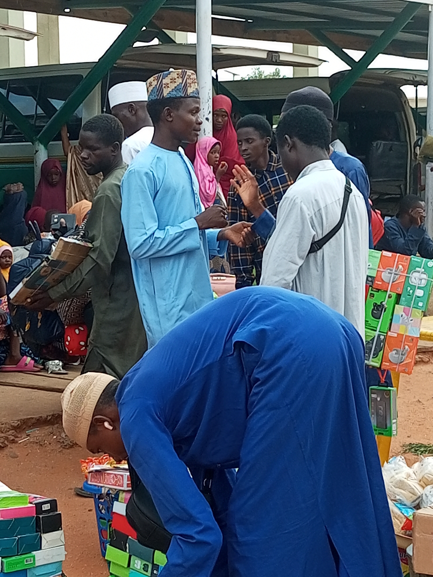In Gombe, Nigeria, a busy street market scene unfolds. A person in a blue robe bends over a cart filled with various items, including boxes and what appear to be packaged goods. They wear a beige skullcap and an earbud. To their right, a display rack holds colorful boxes, likely containing electronics or accessories. In the background, several individuals are gathered, some wearing traditional Islamic attire such as caps and hijabs. Vehicles, including a green bus or van, are visible, suggesting a transportation hub or busy thoroughfare. The ground is sandy and dusty, typical of the region. The atmosphere suggests daily commerce and social interaction in a public space.