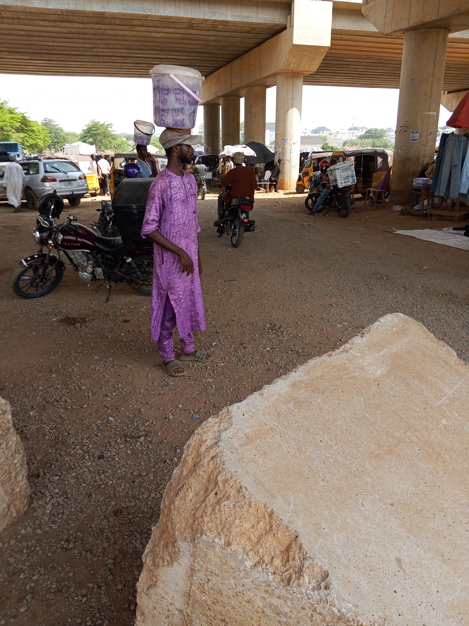 The image depicts a scene in Galadimawa Village, Nigeria, under a concrete overpass. A man wearing a purple robe and a cap balances a plastic bucket on his head. Several motorcycles are parked nearby, and other people are present in the background, along with cars, auto rickshaws, and vendors selling goods. The ground is dirt with loose gravel, and a large block of tan concrete occupies a significant portion of the foreground.