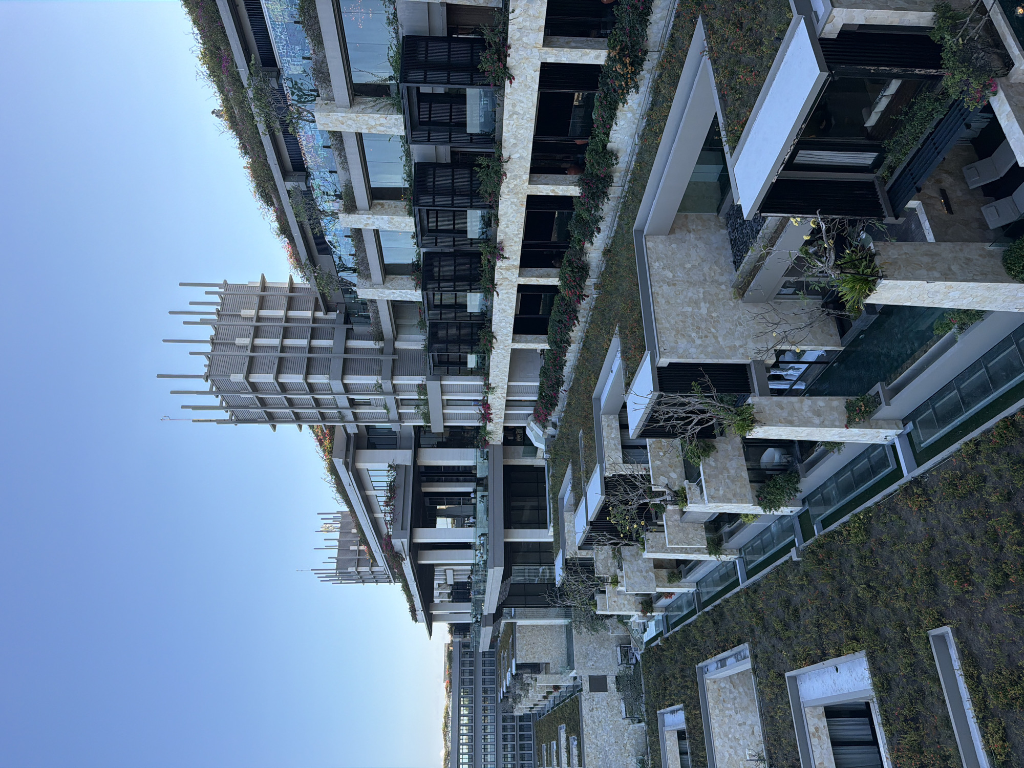 This image showcases a sprawling, modern architectural complex, likely a luxury resort or residential building, situated in Kuta Selatan, Indonesia. The photograph is taken from an elevated perspective, looking across and slightly down at the multi-tiered structures under a clear, bright blue sky.
The complex is characterized by its contemporary design, featuring light-colored stone cladding, concrete elements, dark window frames, and glass railings on balconies. A prominent detail is the extensive integration of greenery; lush vines and flowering plants, including pink and purple blooms, cascade over balconies and adorn numerous building facades, creating a vibrant vertical garden effect. Many flat rooftops and terraced areas are also covered in dense green foliage.
Individual units or sections within the complex feature private outdoor spaces, some with visible water features such as pools, and outdoor lounge areas furnished with sun loungers. Distinctive architectural details include slatted structures atop certain building sections, possibly serving as pergolas or decorative elements. The overall aesthetic suggests a blend of modern luxury and harmonious integration with nature.
The clear, vibrant blue sky indicates a sunny day, likely during the morning or early afternoon. The weather appears calm and pleasant. No people or specific activities are visible within the frame, contributing to a serene and tranquil atmosphere. There is no visible text present in the image.