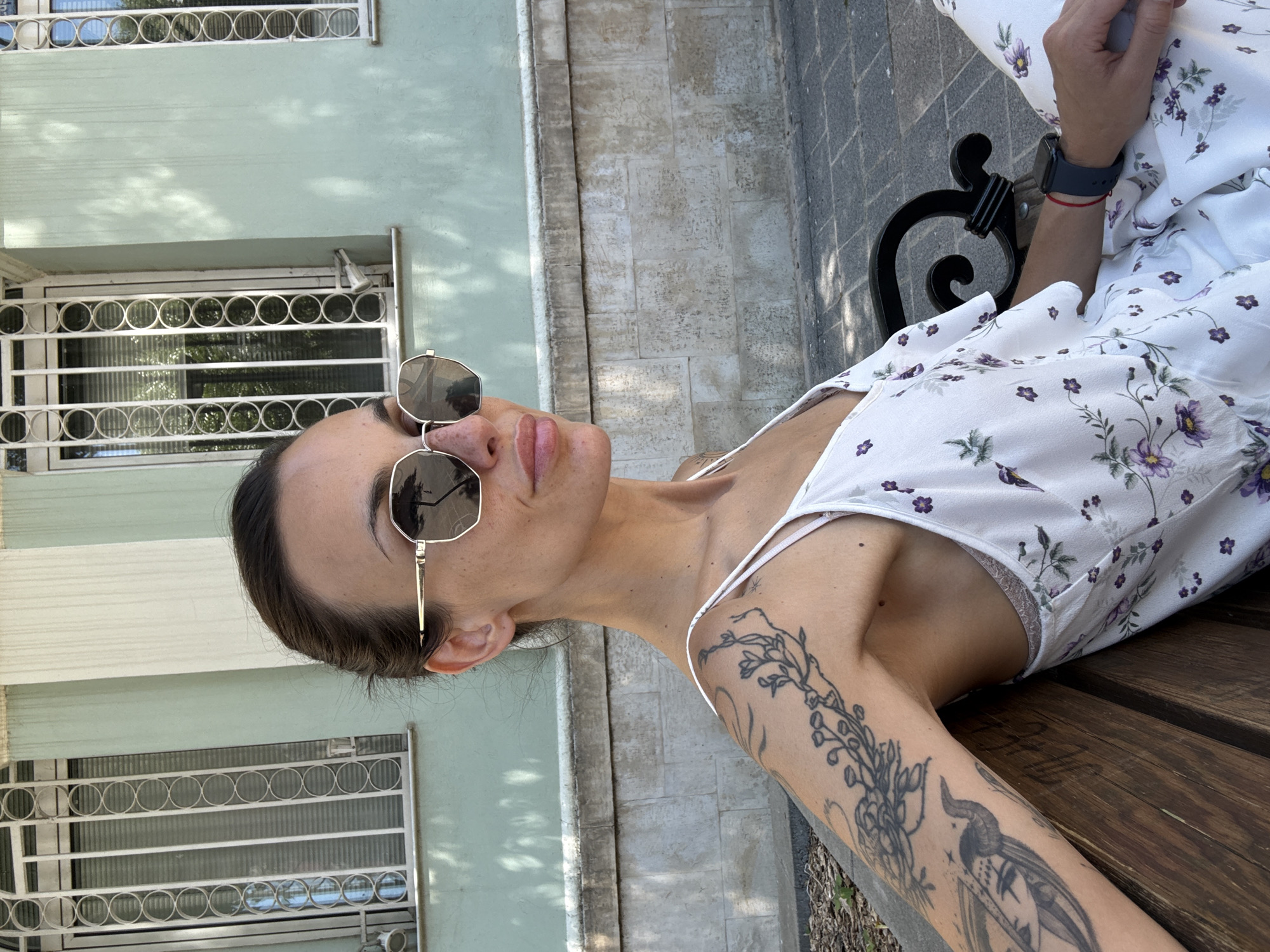 A medium shot captures a woman with dark hair pulled back, wearing sunglasses and a white floral print dress, sitting on a wooden bench. Her left arm, adorned with tattoos, rests on her leg. She is looking to her right, with her mouth slightly open. The background features a light green building with ornate white window grates and a stone wall. The scene appears to be outdoors, possibly in a park or public square, consistent with locations in Pleven, Bulgaria. The lighting suggests it is daytime, with dappled sunlight casting shadows on the building.
