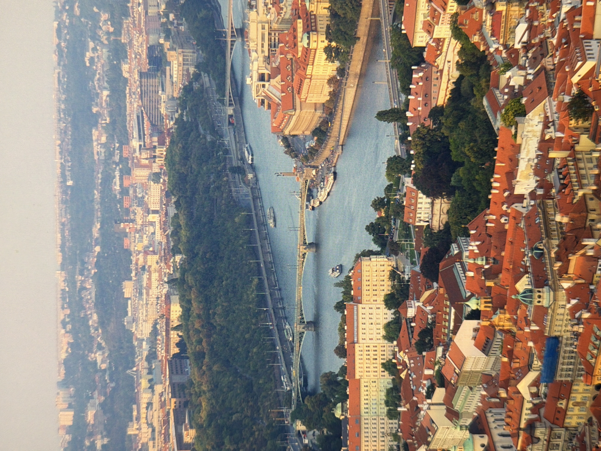 This is a high-angle, wide shot of the city of Prague, Czech Republic, with a focus on its Vltava River and historical architecture. The image captures a sprawling cityscape with numerous red-tiled rooftops, characteristic of Prague's old town. Multiple bridges span the wide, calm river, with boats visible on its surface. The cityscape extends into the distance, with buildings nestled amongst lush green trees and rolling hills. The sky is clear and pale, suggesting a daytime scene, possibly during the late afternoon given the warm lighting on some buildings. The overall impression is one of a historic European capital city.