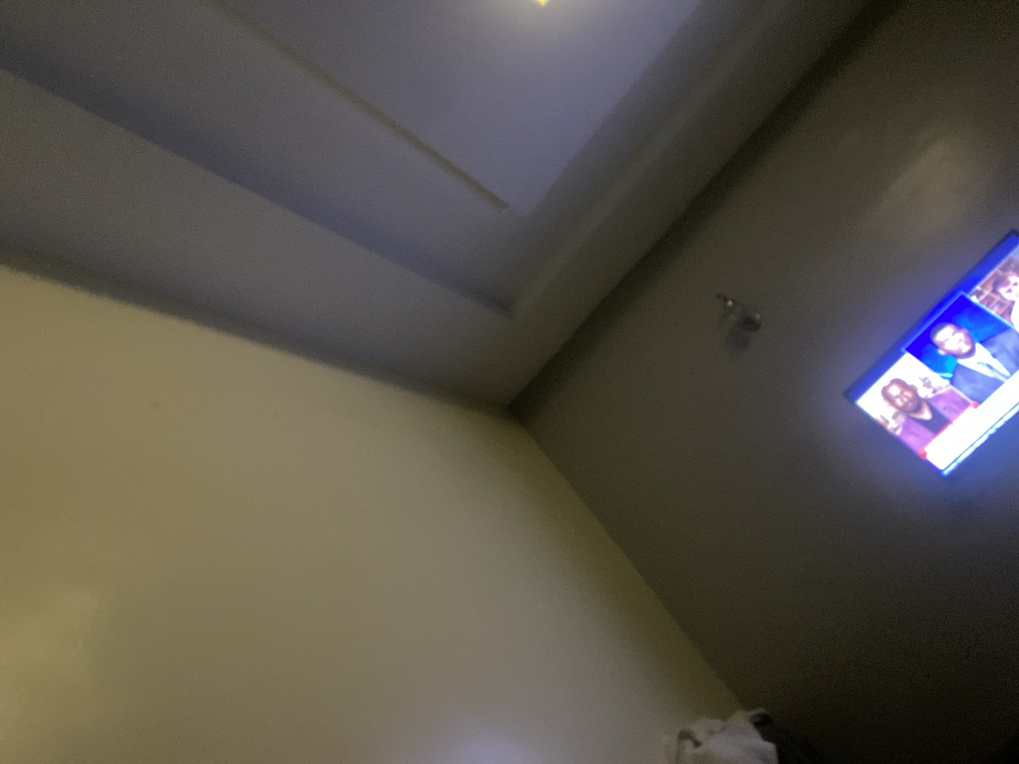 The image displays a dimly lit room with a television screen visible in the upper right corner. The television shows a news program featuring several individuals, one of whom is wearing a purple jacket. The walls are a neutral color, and the ceiling appears to have a layered design. The scene is located in Barikin Jirga Summa, Nigeria. There is no clear indication of any specific actions or interactions taking place.