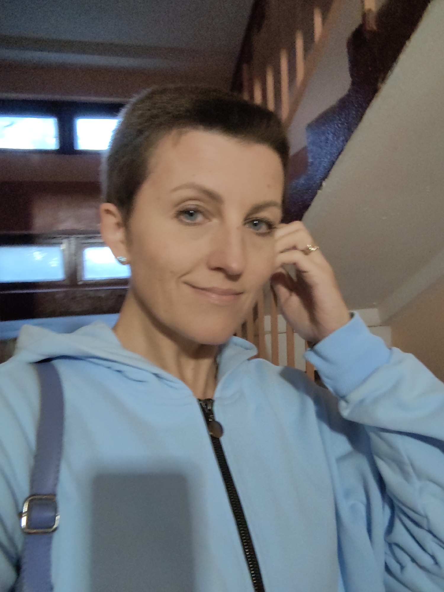 A woman with short brown hair and blue eyes, wearing a light blue zip-up hoodie and a light blue strap across her chest, poses for a selfie. She is wearing a ring on her right hand and a small earring in her left ear. The background appears to be a stairwell with wooden banisters and several square windows. The image was taken in Fanipal, Belarus.
