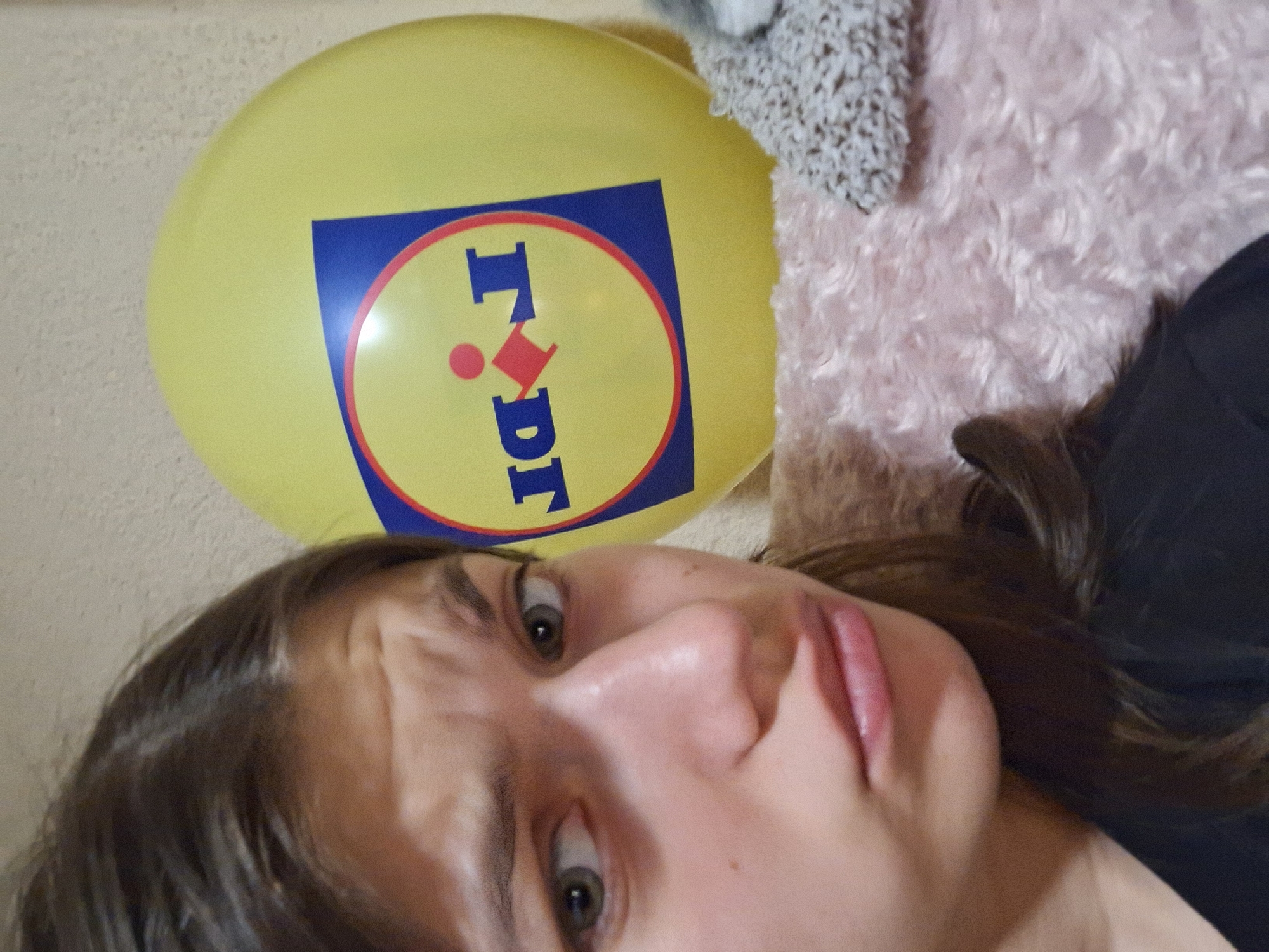A selfie shows a young woman with shoulder-length brown hair and green eyes looking at the camera with a slightly furrowed brow. Above her head to the left is a yellow balloon with the blue and red Lidl logo. Part of a fluffy, light-pink object is visible behind the balloon to the right, along with a textured, light-grey item. The background to the left is a plain, light-colored wall. The woman is wearing a dark-colored top on the right. The location in Panagyurishte, Bulgaria, cannot be determined from the visual information.
