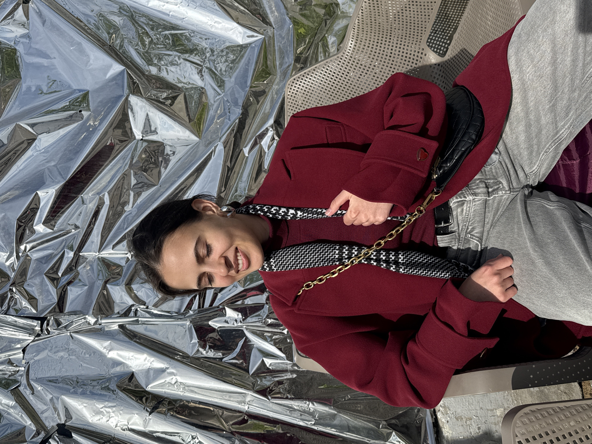 A woman is lying sideways in a gray metal lawn chair in what appears to be an outdoor setting. She is resting on a sheet of crumpled silver foil and is dressed in a dark red coat, gray jeans, and a dark-colored turtleneck sweater. A black purse with a gold chain is slung across her body. The environment is bright and appears to be outdoors, with shadows from trees or other objects falling on the metal chair. The image provides no definitive clues to confirm the location is in Saint Petersburg, Russia, so I am unable to confirm that detail.