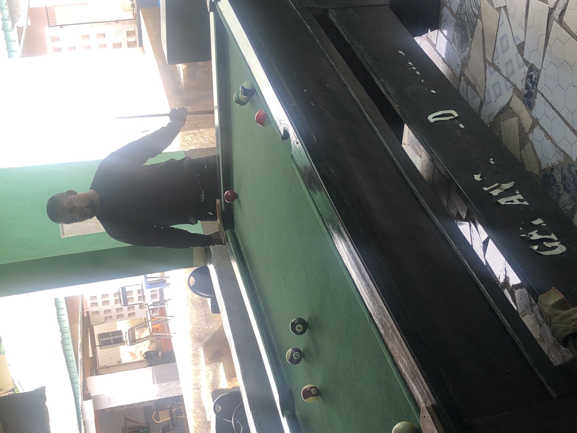 A medium shot captures a Black man standing behind a pool table in what appears to be an outdoor or semi-outdoor establishment in an unknown city. The man is wearing a dark long-sleeved shirt and dark pants, and he is holding a pool cue. The pool table is green, with several balls on it, including a striped ball with the number 5, a solid ball with the number 8, and a striped ball with the number 12. In the background, there is a light green wall, some buildings with windows, and a roof. The ground is tiled. The lighting suggests it is daytime.
