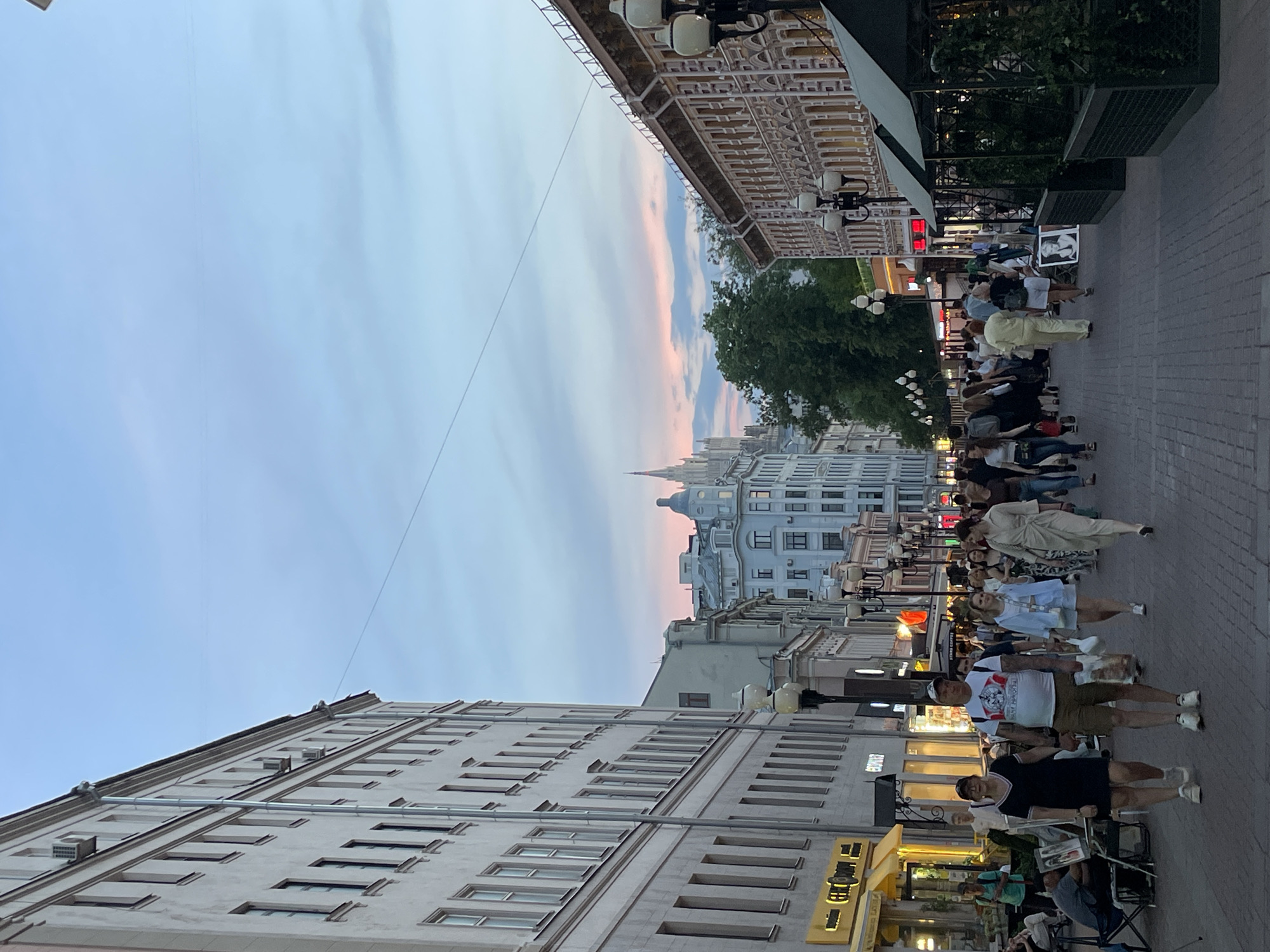 The image shows a street in Moscow, Russia, at dusk. A group of people are standing in a line on the sidewalk, possibly waiting. Buildings line both sides of the street, with one building on the left being a light gray, multi-story building with many windows. The building on the right has ornate architecture and light fixtures. The sky is visible above the buildings, showing a mix of blue and pink hues. The photograph is taken at street level, with a slight upward angle.
