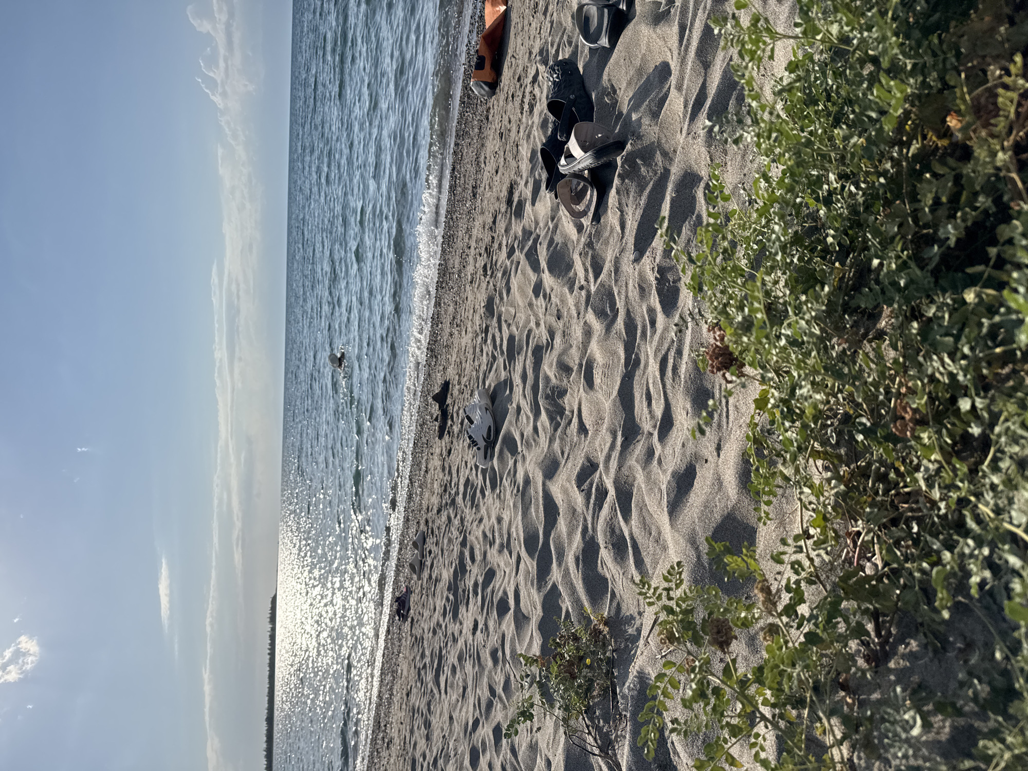 An aerial view looking down onto a sandy beach with the sun glinting off the surface of the blue water of Lake Issyk-Kul, near Basbeshik, Kyrgyzstan. Several pairs of sandals and flip-flops are scattered on the sand, along with an orange inflatable object. A person is swimming in the distance. The beach is sparsely vegetated with a few small green plants.