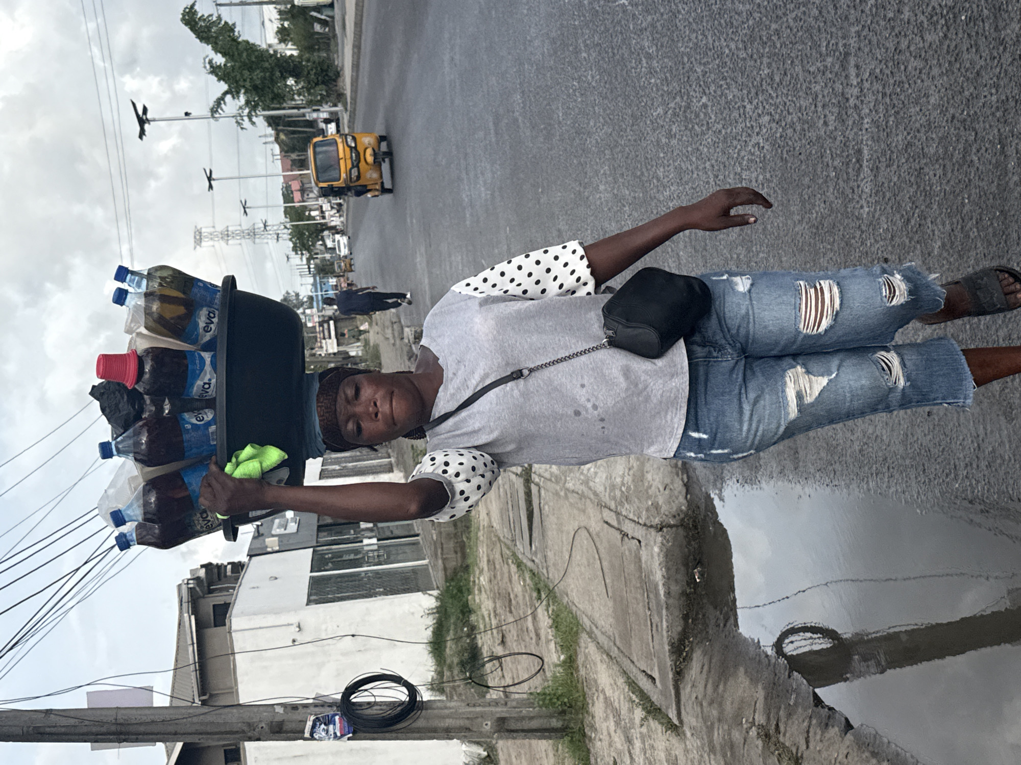 A woman walks along the side of a paved road in Ado, Nigeria. She carries a large black basin filled with bottles of water and other drinks on her head. She wears ripped jeans, a gray shirt with polka-dot sleeves, and sandals. A small black bag hangs from her shoulder. A yellow tricycle-type vehicle is visible on the road in the background, along with other distant cars and buildings. Power lines are overhead. A puddle of water reflects the sky and a portion of a nearby building.