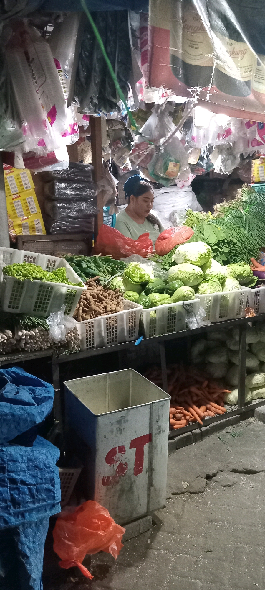 The image depicts a street-side market stall in Babat, Indonesia. A woman with dark hair pulled back in a bun is seated behind a display of fresh produce. She is wearing a light green shirt and appears to be attending to the vegetables. The stall is laden with various fruits and vegetables, including lettuce, cabbage, green beans, chilis, carrots, and tamarind, displayed in white plastic crates. Behind the woman, there are stacks of black plastic bags and some yellow packaged goods on shelves. The stall is covered by a tarpaulin, and various plastic bags and other items are hanging from the structure. In the foreground, there is a large blue tarp, a weathered metal trash bin with red markings, and a bright orange plastic bag. The ground appears to be paved and somewhat uneven, with some debris scattered on it. The lighting suggests it might be evening or a dimly lit area. The overall scene conveys the typical atmosphere of a local market in Indonesia.