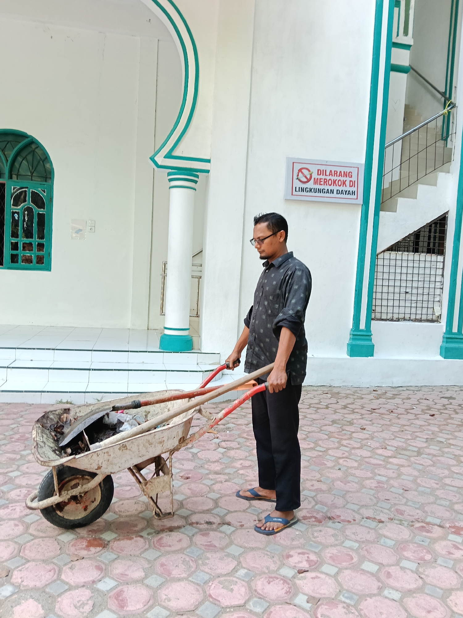 An outdoor, eye-level, full-shot photograph captures a man standing beside a wheelbarrow filled with debris, suggesting a cleaning activity. The scene is set on a paved area with red and grey hexagonal tiles, next to a white building featuring distinctive green architectural accents, including arched windows, columns, and decorative trim. A concrete staircase with metal railings is visible on the right side of the building.
The man, positioned slightly left of center, is of medium build, wearing a dark patterned long-sleeved shirt, dark trousers, and blue flip-flops. He has short dark hair, a beard, and glasses, and he is holding the red-handled wheelbarrow, which appears aged and contains what looks like dirt, a shovel, and other discarded items. His gaze is directed downwards, focused on the wheelbarrow or the task at hand.
The building's white walls and green details, including an ornate archway, suggest a religious or educational institution, which is confirmed by a prominent red and white sign on the wall. The sign clearly reads "DILARANG MEROKOK DI LINGKUNGAN DAYAH," translating to "NO SMOKING IN THE DAYAH ENVIRONMENT." A 'dayah' is an Islamic boarding school in Indonesia. The environment appears bright, indicating daytime. The image was taken in Lhokseumawe, Indonesia.