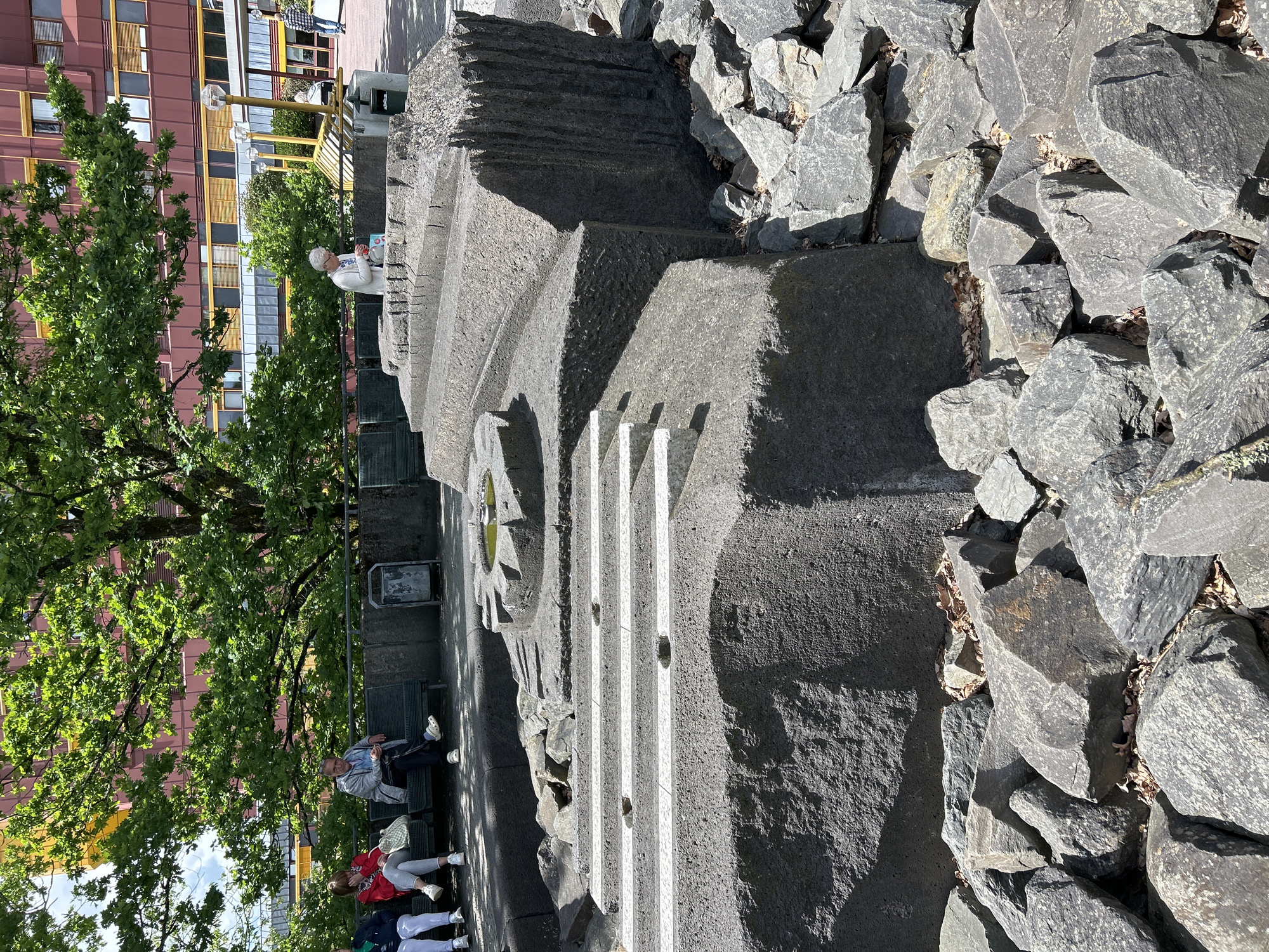The image depicts an outdoor public space in Gummersbach, Germany, on a clear, sunny day. In the immediate foreground and extending into the midground is a large, dark grey stone sculpture or installation. This structure is composed of multiple geometric elements, including a substantial rough-hewn block base, a section with three horizontal light-colored (possibly white) parallel lines or steps, and an elevated circular basin containing stagnant greenish water, suggestive of a non-operational fountain or pool. The right side of the primary stone mass exhibits vertical striations or grooves. Surrounding the base of this stone feature is an irregular arrangement of smaller, dark grey rocks and scattered dry leaves.
In the midground, behind the stone structure, a row of dark-colored park benches is visible. On the left bench, two individuals are seated. The leftmost person is partially visible, wearing a dark top and light pants. The person next to them, centrally positioned, wears a red long-sleeved top, light grey pants, and white sneakers, with a light-colored handbag beside them. On an adjacent bench to the right, a man is seated, wearing a light grey zip-up jacket over a blue shirt, dark pants, and light-colored slip-on shoes, engrossed with a small electronic device. Further to the right, a woman in a white cardigan and light pants is standing or leaning over the stone structure, appearing to interact with an item in her hands near the circular basin. A dark grey public trash bin is positioned between the seated groups.
The background features a large deciduous tree with dense, vibrant green foliage, casting dappled shadows on the ground. Through and above the tree canopy, a multi-story building with a reddish-brown facade and numerous windows, some fitted with yellow or orange sun shades, is visible. Several yellow, multi-pronged decorative streetlights or fixtures are present on the right side of the background. A person wearing a plaid shirt is discernible walking in the far-right background. The sky is partially visible, appearing light blue with scattered white clouds.