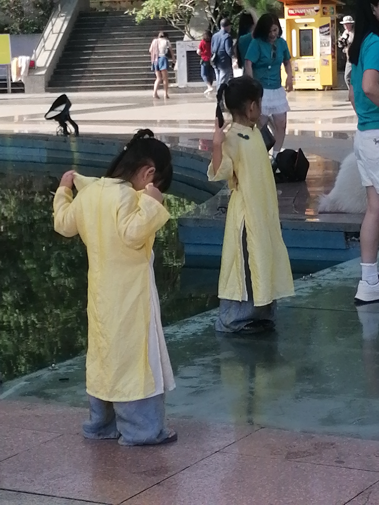 The image depicts an outdoor scene, possibly in a public space with a water feature. In the foreground, two young girls are visible. The girl on the left is wearing a yellow, long-sleeved garment over grey pants and appears to be looking down. The girl on the right is also wearing a similar yellow garment, the sleeves of which are pulled up, and she is looking towards the left. Behind them, several adults are present. One woman in a turquoise shirt and white skirt is visible, along with other figures in the background. A yellow vending machine is also present. The environment includes a tiled ground surface and a body of water, possibly a pool or fountain. A black bird-shaped object is also seen near the water's edge. The overall lighting suggests it is daytime. There is no information in the image to place it specifically in Da Lat, Vietnam.