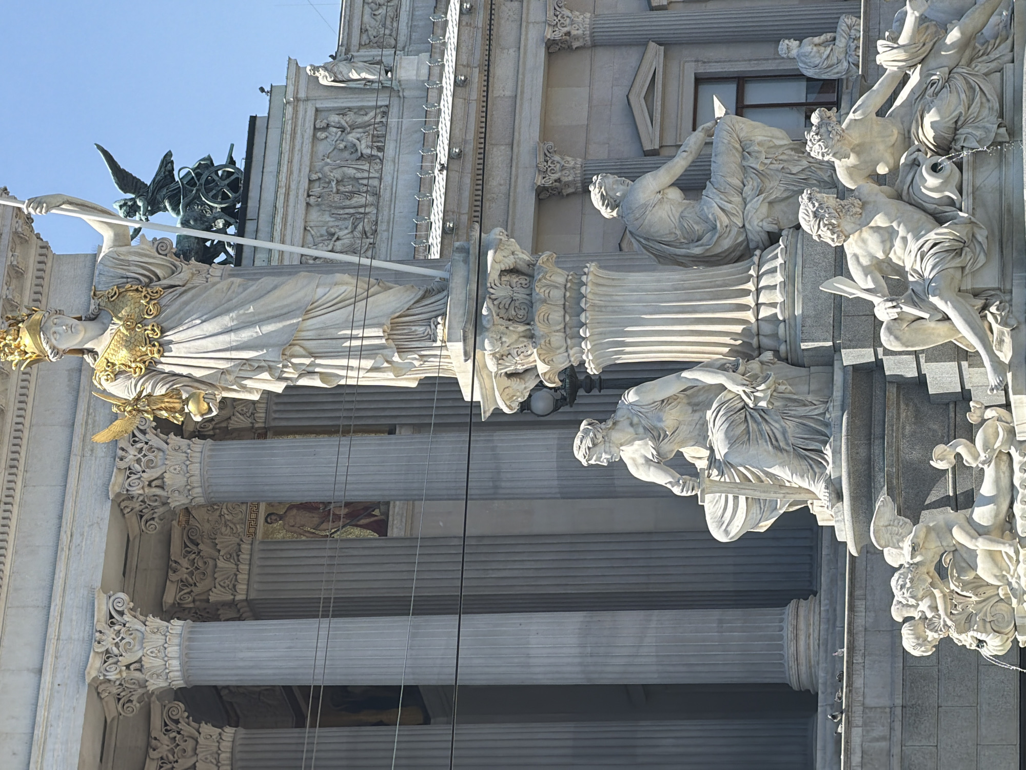 This is a photograph of the Pallas Athene Fountain in front of the Austrian Parliament Building in Vienna, Austria. The fountain features a large central statue of Pallas Athene, the Greek goddess of wisdom and war, holding a spear and a golden orb. She is adorned with a golden breastplate and a crown. At her feet are sculptures of figures from mythology and allegorical representations. In the background, the grand neoclassical facade of the Parliament Building is visible, characterized by its numerous columns and ornate architectural details. The sky is clear and blue, indicating a sunny day. The scene captures a public monument and historical architecture in an urban setting.