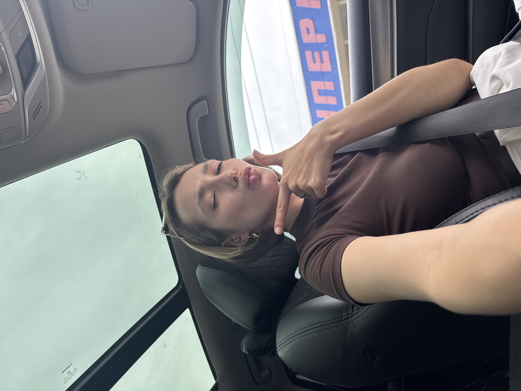 A young woman with short blonde hair sits in the passenger seat of a car, her eyes closed and lips pursed as she makes a kissing gesture. She is wearing a brown t-shirt and a silver ring. She has a gold heart-shaped earring in her left ear. Her right hand is raised, with her thumb and index finger extended, pointing towards the side. The car's interior is visible, including black leather seats and a sunroof. Outside the car, a building with large blue and pink lettering is visible. The overall scene appears to be captured during daylight.