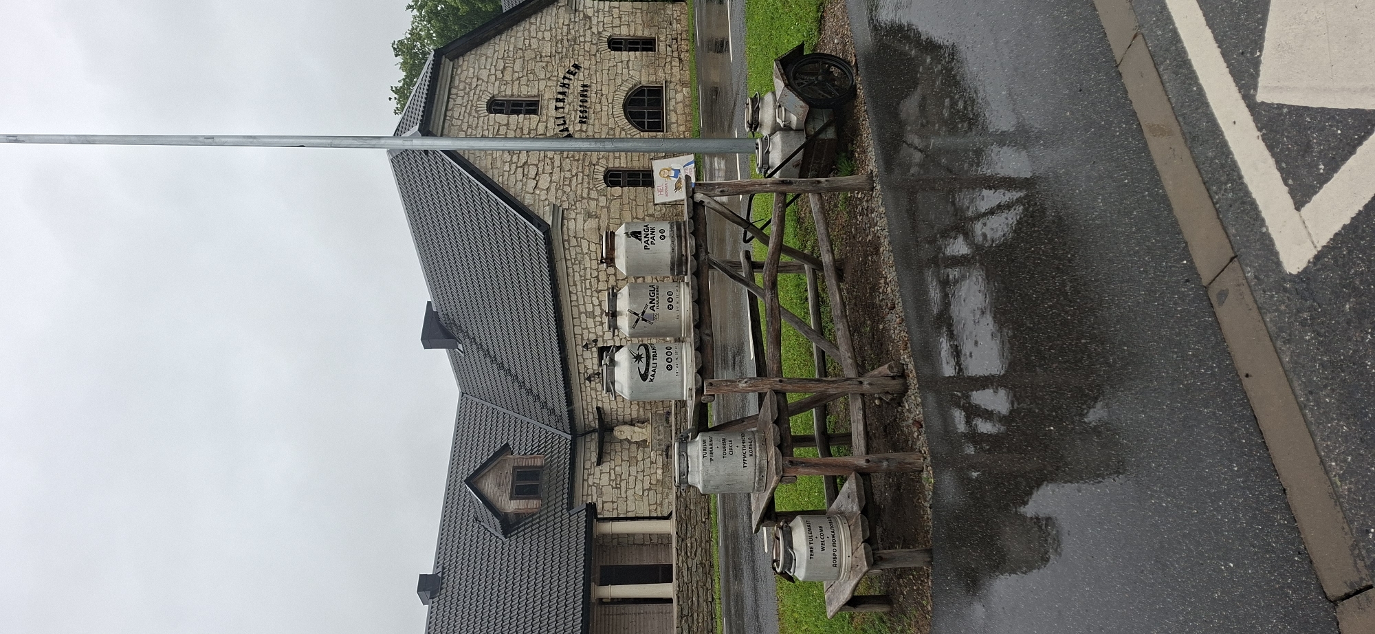 In Kaali, Estonia, an overcast sky looms over a stone building with a dark, tiled roof. The building, identified by signage as "KAALI TRAHTER RESTORAN", features arched windows and a dormer window. In front of the building, a rustic wooden table is set up, displaying several large metal milk churns. Three churns are placed on the table, each with labels indicating "KAALI TRAHTER", "ANGLA", and "PANGA PANK". To the left, another churn sits on a wooden step, labeled "TURISM 'PIIMARING'". A smaller churn is placed on a lower wooden step and is labeled "TERE TULEMAST WELCOME ДОБРО ПОЖАЛОВАТЬ". A small, wheeled cart with two more churns is positioned to the right of the table. The ground is wet and reflective, indicating recent rain. The scene appears to be set up as a display or welcome area for visitors.