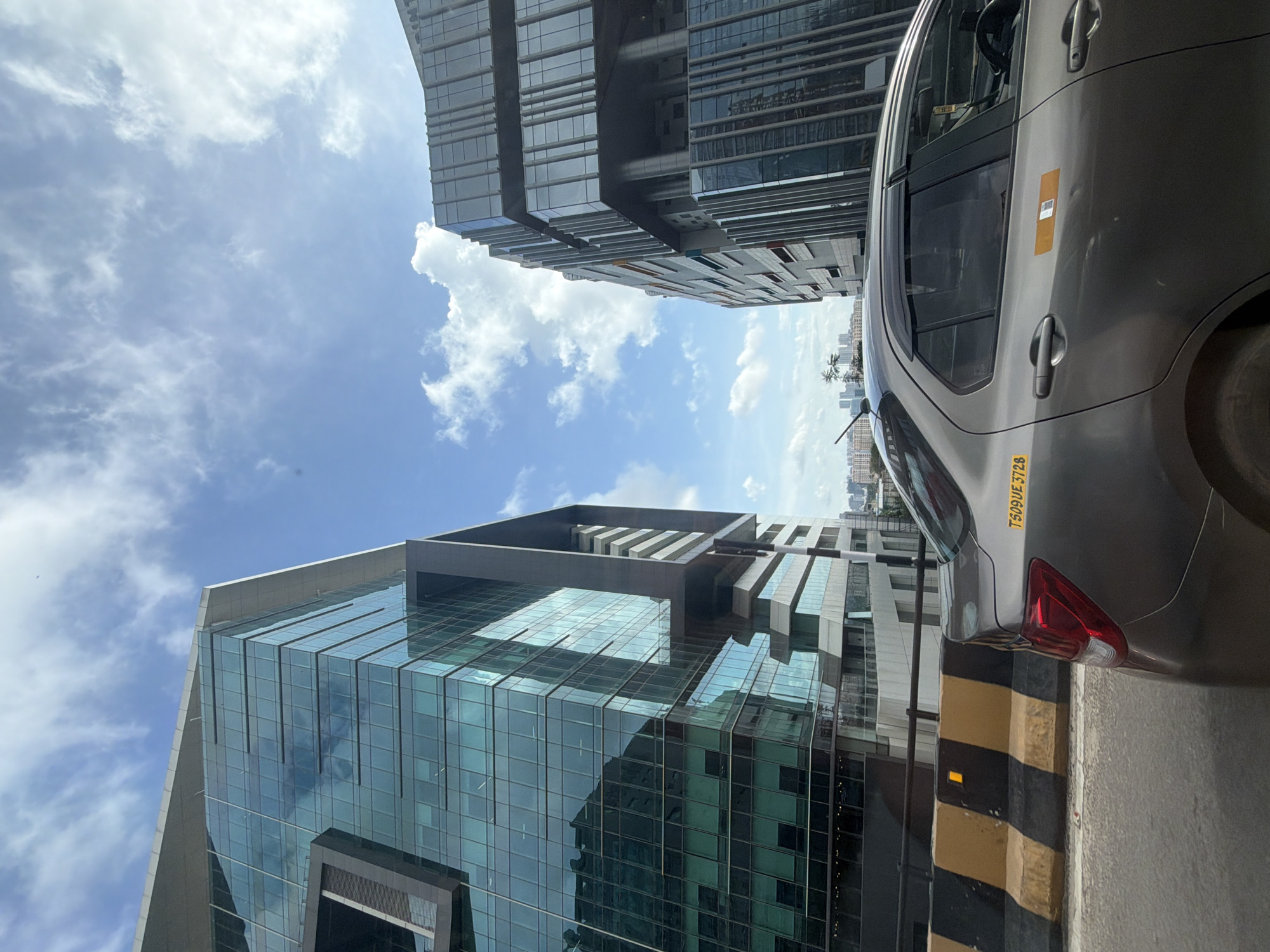 The image captures a section of the HITEC City area in Hyderabad, India, under a partly cloudy sky. Two modern, glass-facade buildings dominate the background, one on the left and another on the right, with the one on the left appearing taller and featuring a distinctive angular design. In the foreground, a gray sedan is traveling on a road. The car has a yellow sticker on its rear fender with the text "TS09UE3728". A yellow and black striped barrier is visible along the side of the road. The overall scene suggests an urban environment with significant commercial development.