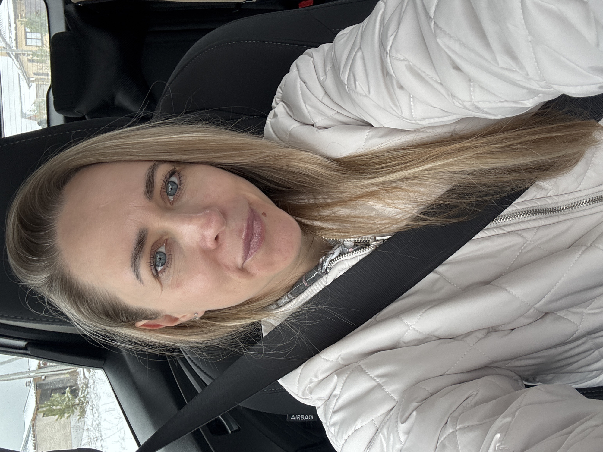 A woman with blonde hair and blue eyes is in a car wearing a white quilted jacket and a seatbelt. The location appears to be Chaikovsky, Russia, based on the buildings and wintery conditions visible through the car window. The focus is on the woman's face. The scene is indoors, inside the vehicle.