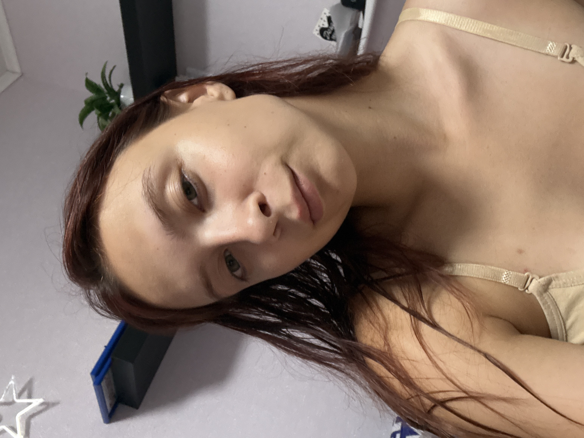 A young woman with long, auburn hair is looking at the camera in a close-up, selfie-style shot. She is wearing a light-colored bra. The background is a pale purple wall with a shelf mounted on it. On the shelf, there is a black object, possibly a book or a drawer, and a small green plant in a white pot. To the left of the shelf, a white star decoration is visible on the wall. The lighting is soft, suggesting an indoor setting. There are no visible elements that can indicate the specific location within Прикамский, Russia.