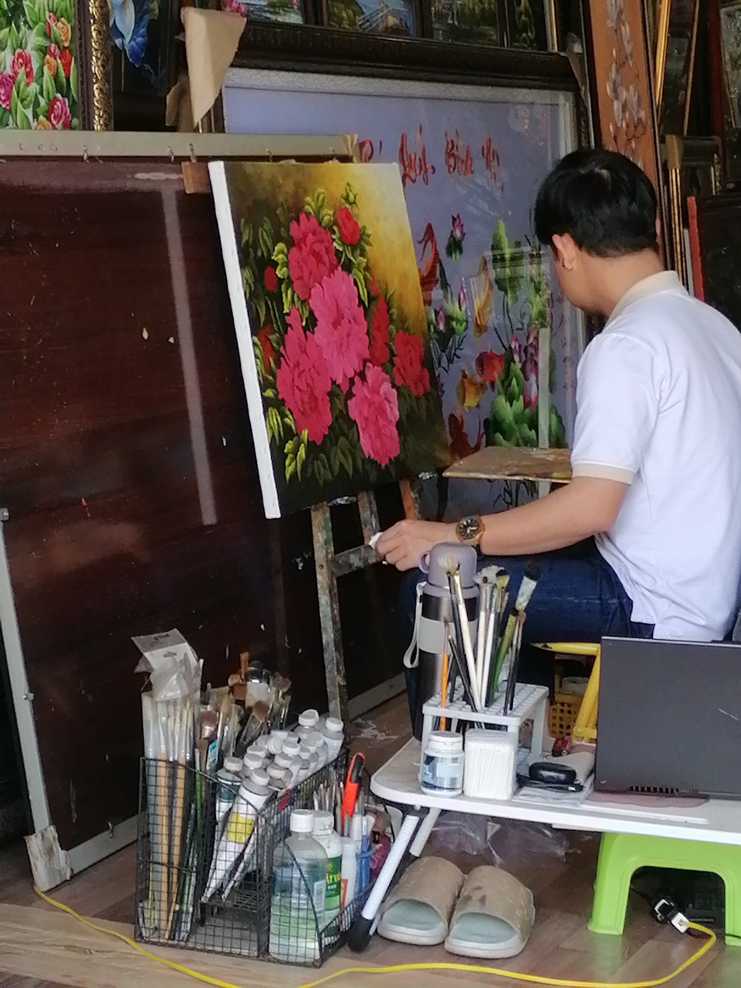 The image depicts an artist working on a painting in a studio, likely located in the Lam Vien - Da Lat area of Vietnam. The artist, a man wearing a white polo shirt and blue jeans, is seen from the side, focused on his canvas. He is holding a brush in his right hand and appears to be actively painting. In front of him, on a low table, are various art supplies, including tubes of paint, brushes in a holder, and what appear to be cotton swabs or applicators. A laptop is also visible on the table. The background shows a collection of framed artworks, suggesting a professional art space or a shop displaying artwork. The overall scene conveys a sense of dedicated artistic creation.