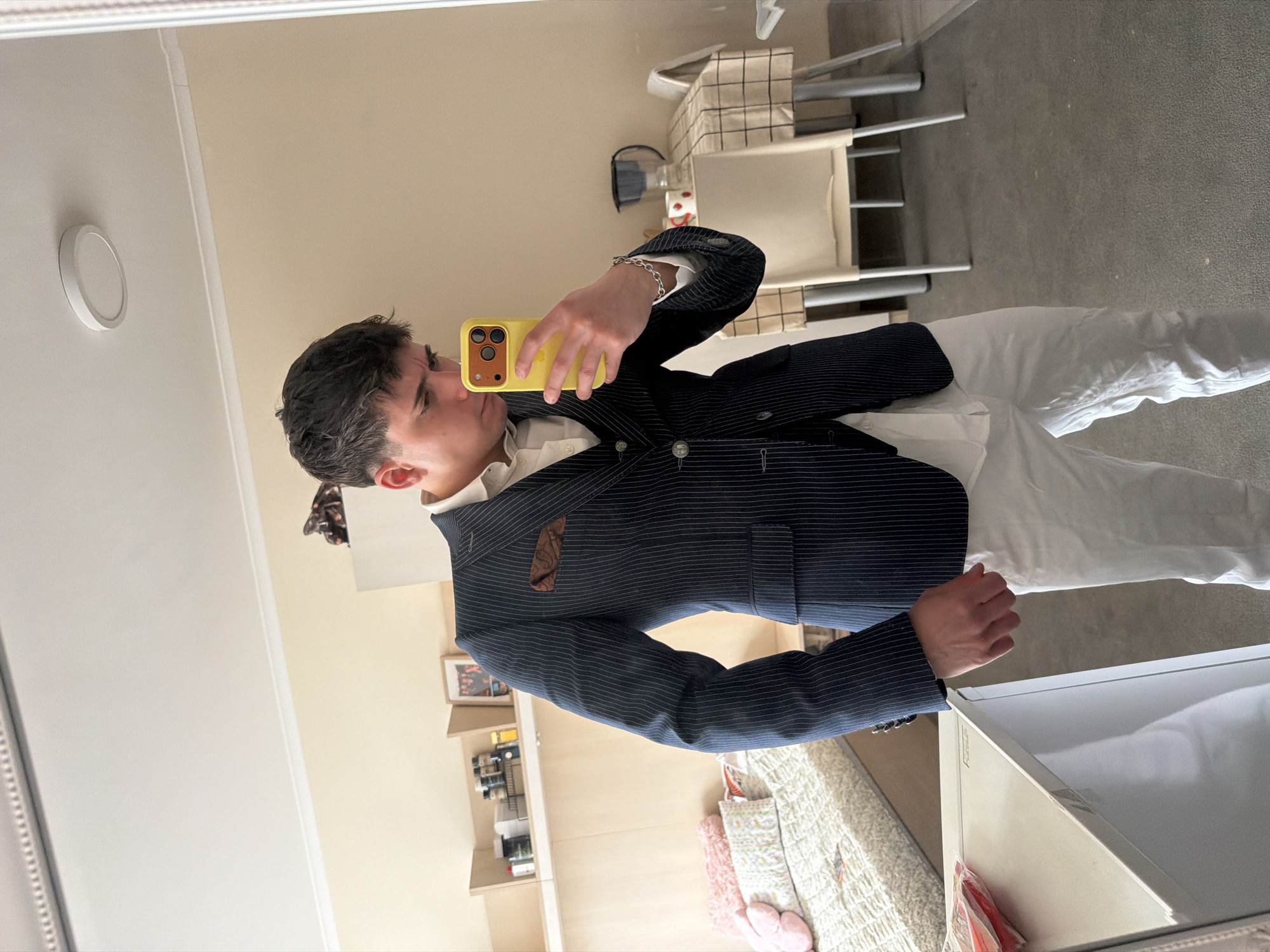 A person takes a mirror selfie in an interior setting. The individual, seen from the waist up, wears a dark, pinstriped blazer over a white shirt and white trousers. They hold a yellow smartphone to capture the image. The background includes a light-colored wall, shelves containing various objects, a bed with a patterned cover, a chair, and a white object with a lid. The location is unknown and there is no evidence that this image was taken in Sofia, Bulgaria.