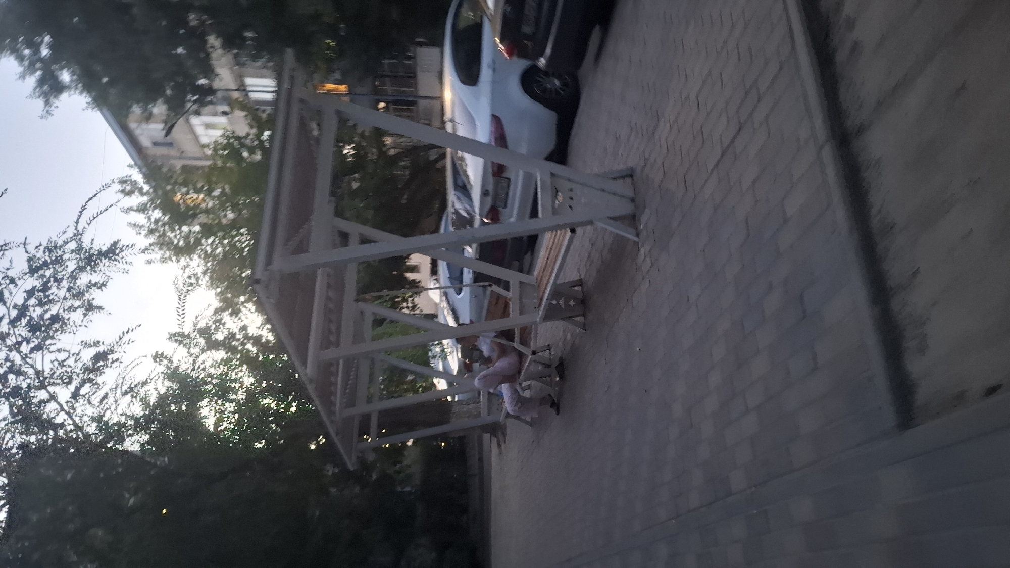 The image depicts a person sitting on a bench under a canopy in a city park area. The person is wearing light-colored clothing and appears to be looking down. Behind the bench, several cars are parked in a row. The environment includes trees, paving stones on the ground, and a building in the background. The lighting suggests it is late afternoon or early evening. The presence of a building with multiple windows and a street lamp suggests an urban setting, possibly residential. The overall scene conveys a sense of calm or repose. This specific scene is located in the city of Shymkent, Kazakhstan, likely on a public pathway or recreational area within the city.