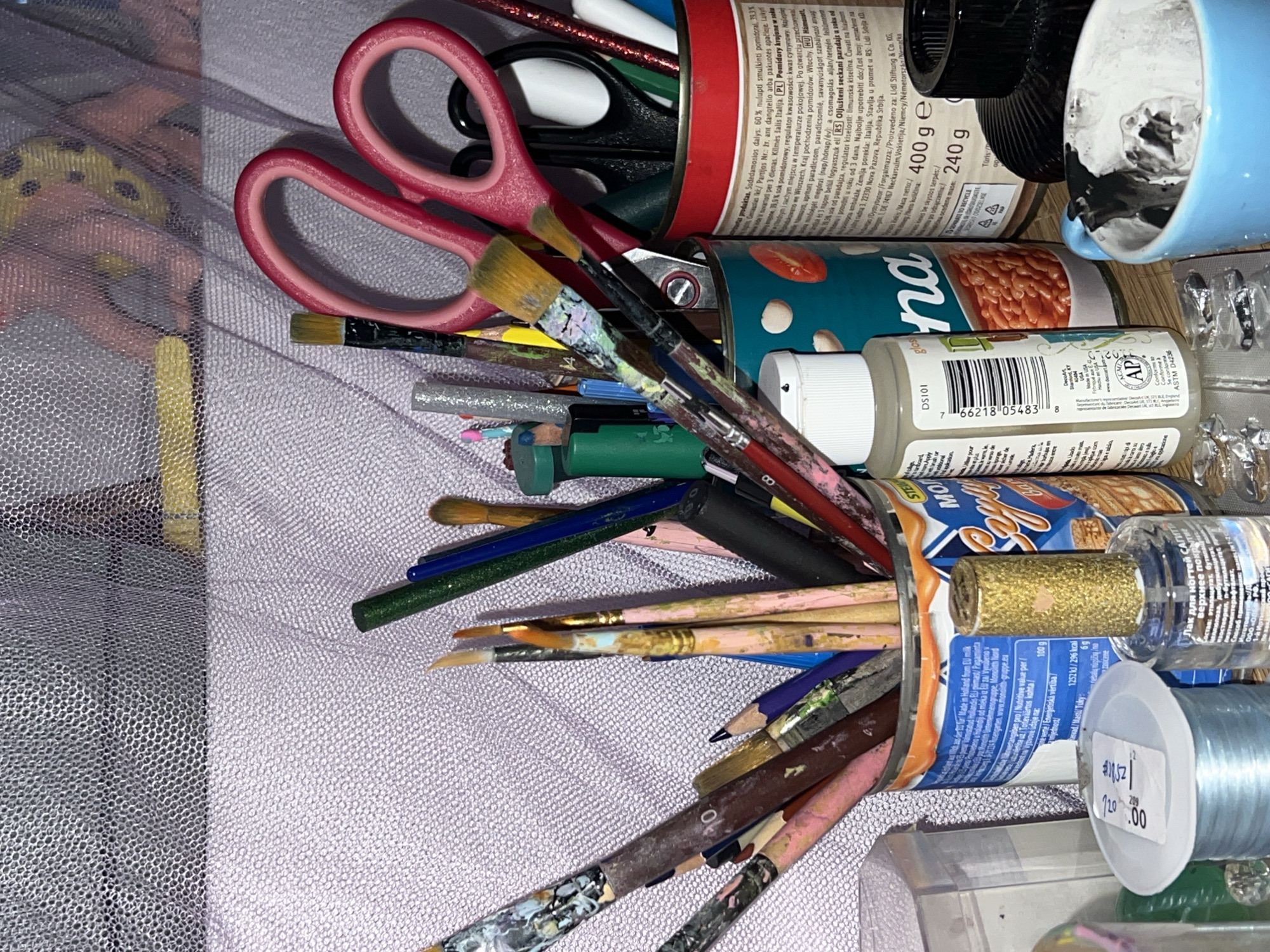 The image presents a chaotic arrangement of art and craft supplies. Various paintbrushes, pencils, and scissors are jumbled together with opened cans used as containers. One can is labeled as chopped tomatoes in tomato juice and one is baked beans. The cans are next to a bottle of nail polish and a roll of string. The supplies are scattered across a lavender fabric surface. A tulle fabric covers the edge. There is nothing in the image to indicate this is in Palilula.