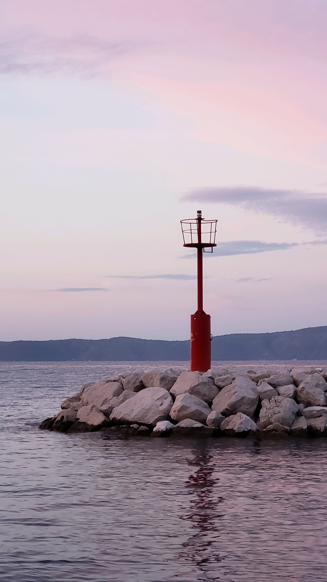 A red lighthouse stands tall on a breakwater made of large, light-colored rocks. The lighthouse has a cylindrical base and a thinner shaft topped with a cage-like structure containing a light. The breakwater extends into calm, dark water that reflects the lighthouse and the sky. In the background, a hazy, mountainous coastline is visible under a soft, pastel-colored sky with wisps of pink and purple clouds, suggesting either dawn or dusk. The scene is serene and isolated, conveying a sense of quiet coastal landscape in Općina Tučepi, Croatia.