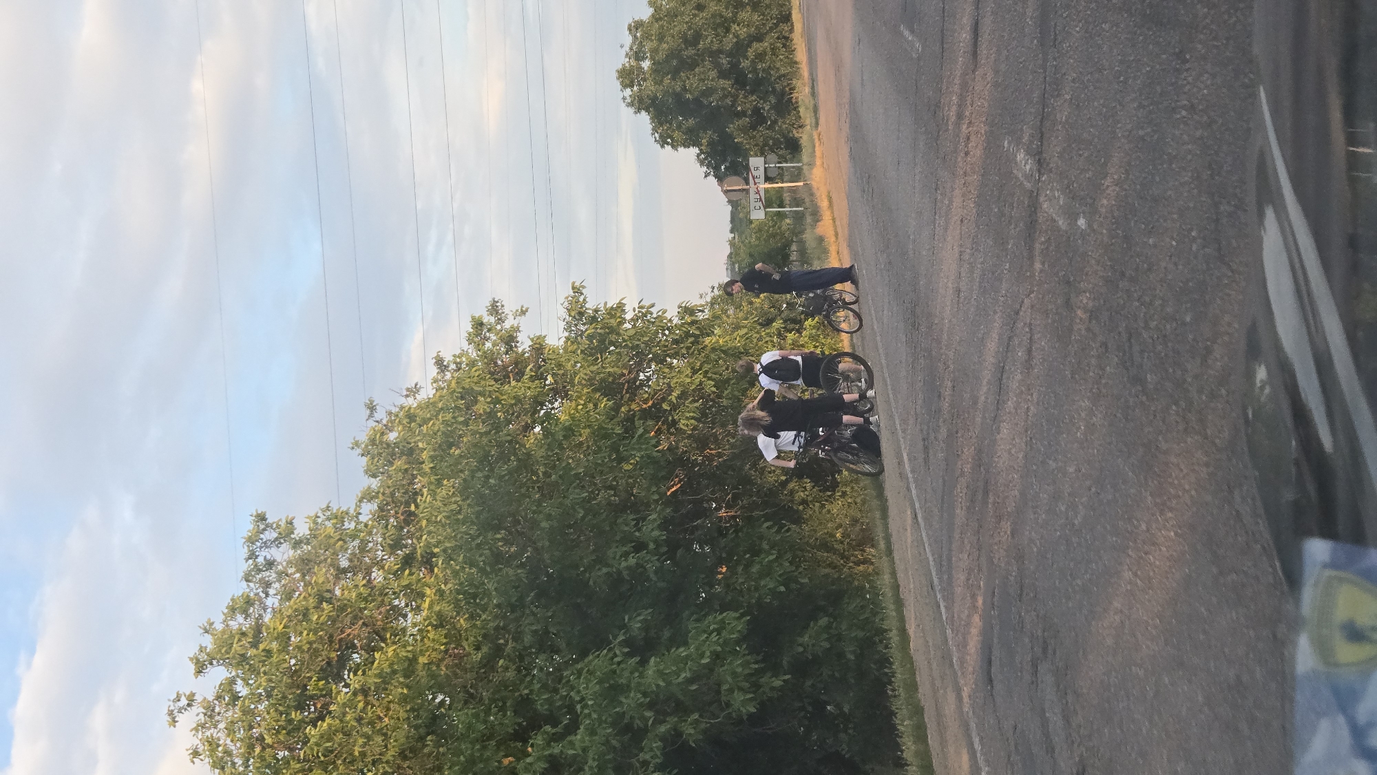 The image depicts a roadside scene in the city of Sukleia, Moldova, with three individuals standing near two bicycles and a motorcycle. A fourth individual, dressed in dark clothing, stands slightly apart. The group appears to be gathered on the shoulder of a paved road. In the background, a sign with Cyrillic lettering is visible, and a tree-lined landscape stretches towards the horizon under a partially cloudy sky. Power lines are strung across the upper portion of the image. The overall atmosphere suggests a pause or gathering during outdoor travel.