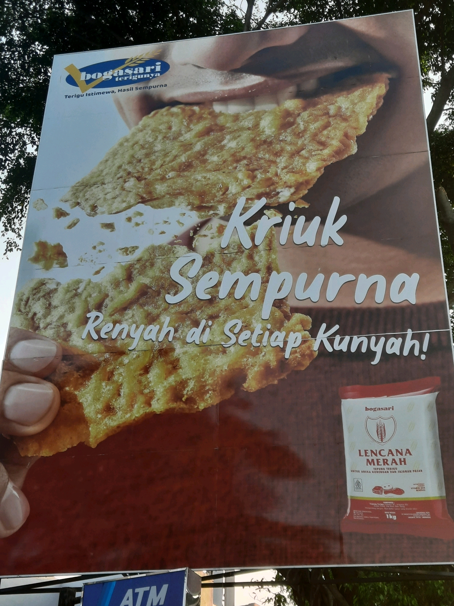 The image shows a billboard advertising Bogasari flour in East Jakarta, Indonesia. The billboard features a close-up shot of a crispy fried food item being held by a person's fingers, with the text "KRIUK SEMPURNA" (Perfectly Crispy) and "Rennyah di Setiap Kunyah!" (Crispy in Every Bite!) prominently displayed. A bag of "Bogasari Lencana Merah" flour is also shown on the right side of the billboard, with a net weight of 1kg indicated. The background shows some greenery, likely trees, which suggests the billboard is outdoors. Below the billboard, a part of an "ATM" sign is visible.