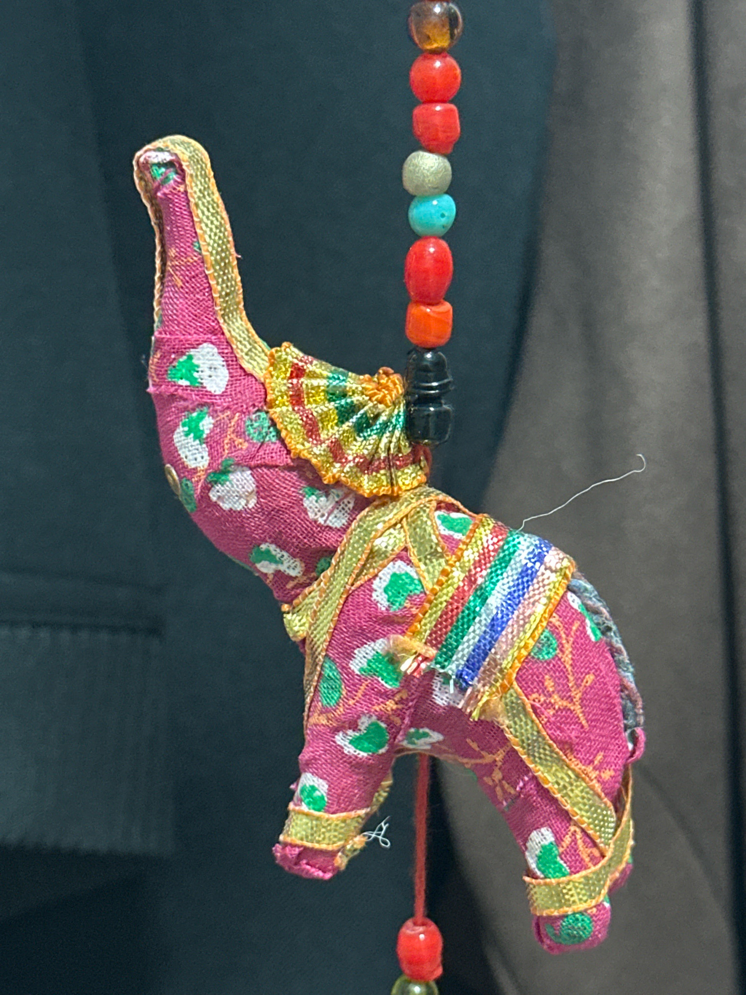 A small, pink fabric elephant figurine hangs suspended by a string. The elephant is adorned with a decorative, colorful fabric on its back and is trimmed with gold ribbon. A string of colorful beads, including red, orange, turquoise, and brown, hangs above the elephant. A white thread is visible near the elephant's back. The background is dark and appears to be a blurred fabric. The image does not provide any information about the location in the city of Nevinnomyssk, Russia.