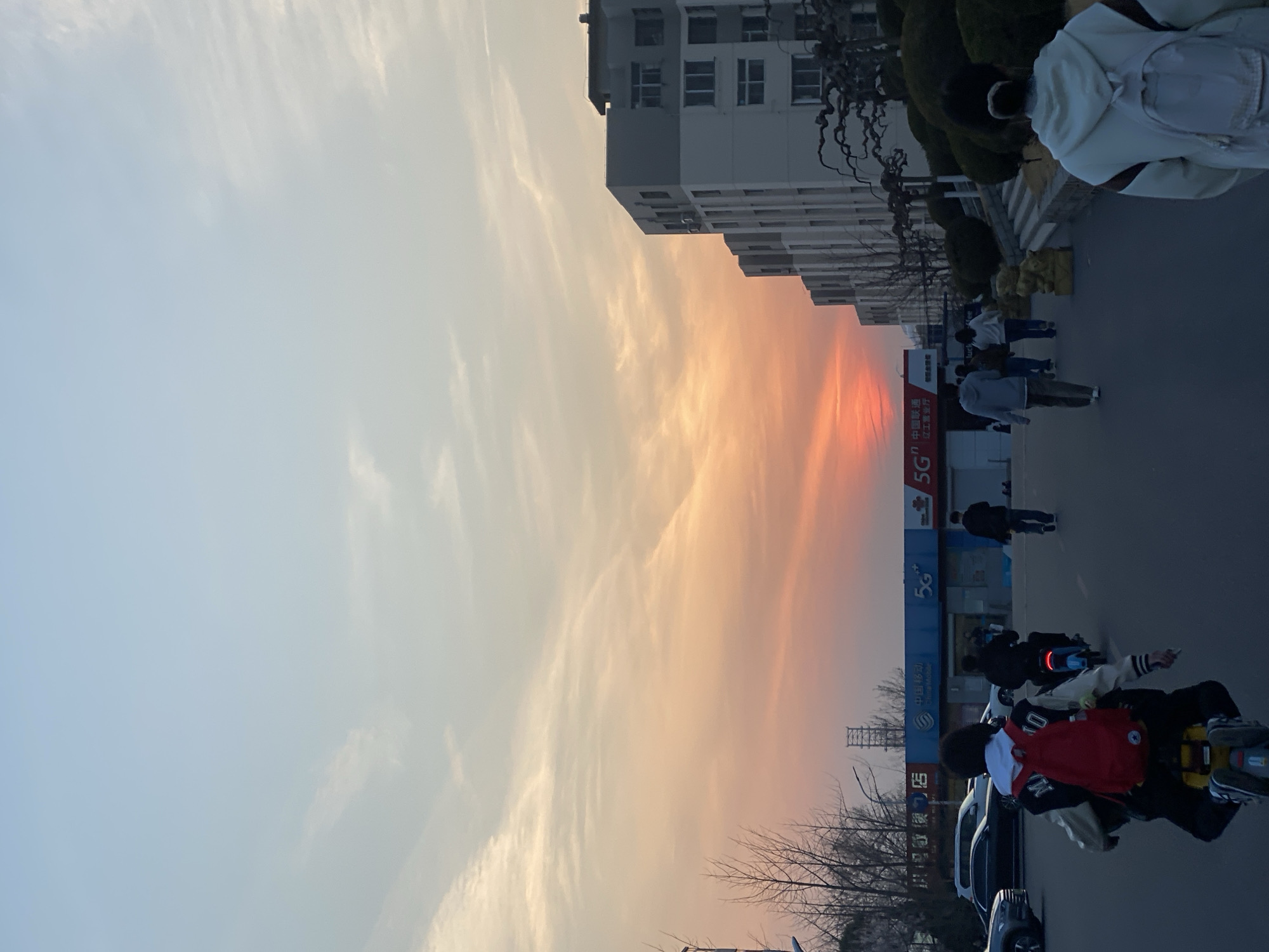 A street scene at sunset features a building, a 5G China Mobile advertisement, and traffic. People are walking on the sidewalk, while others are riding electric bikes in the street. Bare trees are visible along the street. The sky is filled with orange and yellow hues indicating the sunset. Text visible identifies a China Unicom branch near Liaogong University in Zhongtun.