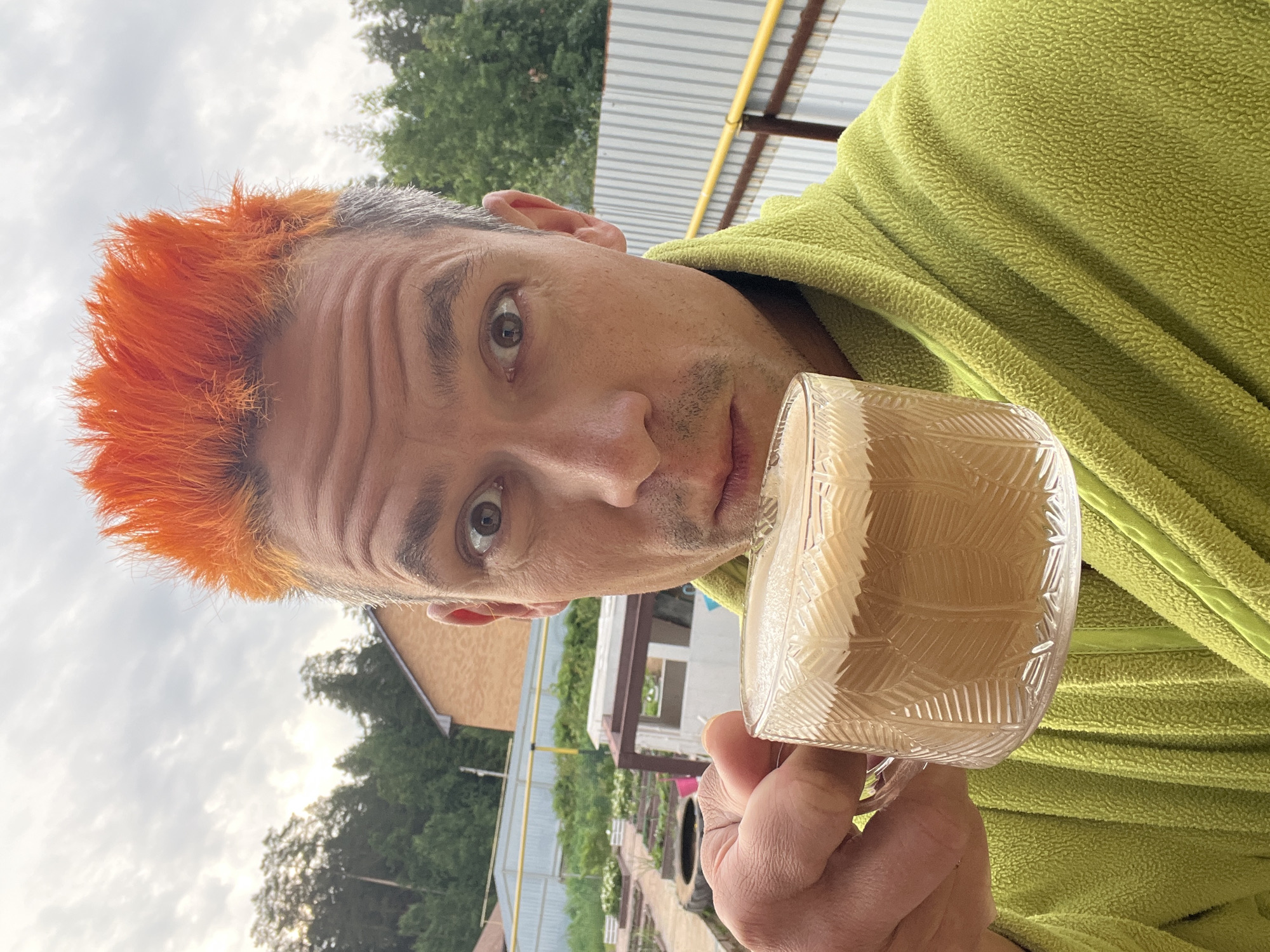 This close-up, vertically oriented image features a man outdoors, likely in a backyard or rural setting in Chaikovsky, Russia. He looks directly at the camera with wide, inquisitive eyes and a slightly surprised expression. His hair is a striking bright orange and spiky at the front, fading into a shorter, greyish tone towards the back and sides. He has a light stubble and visible wrinkles on his forehead.
The man is wearing a lime green, textured fleece jacket or robe. He holds a clear glass mug with an etched, decorative pattern, containing a light brown beverage, possibly coffee or tea with milk or creamer, visible with a slight frothy top.
The background is an outdoor scene under an overcast sky. Visible elements include dense green trees, a fence with metal posts and corrugated panels, a building with light brown siding, and a yellow pipe running horizontally in the upper part of the frame. The setting suggests a casual, possibly semi-rural environment. No visible text is present.