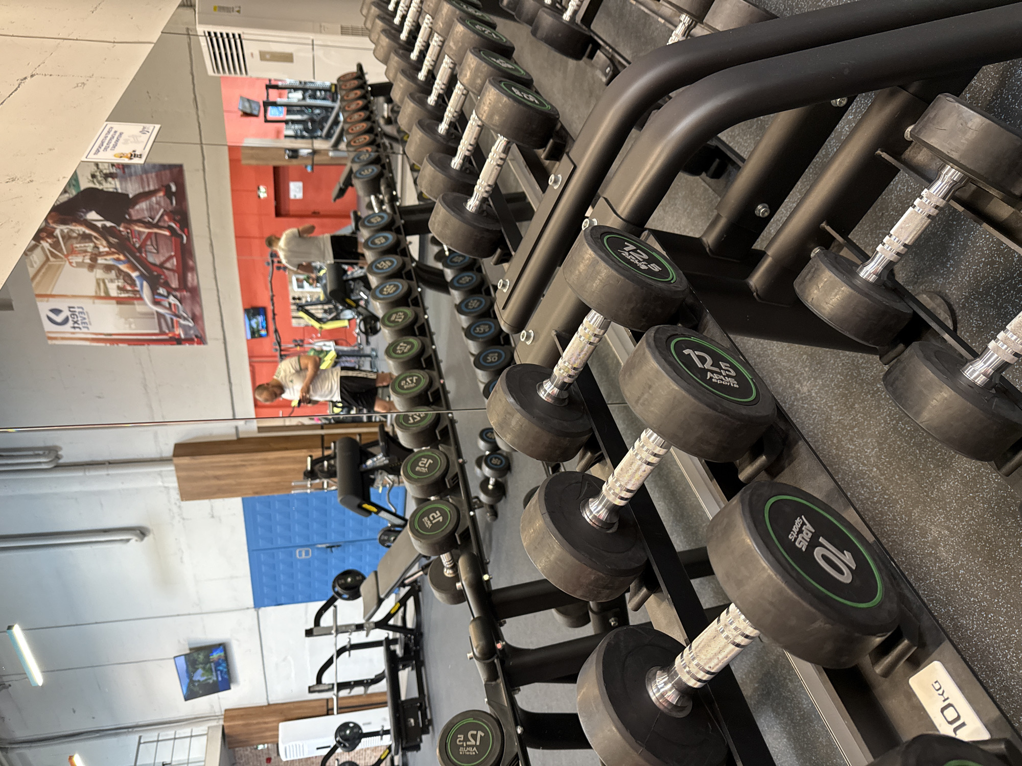 The image captures a weight rack in a gym, likely located in Sofia, Bulgaria given the presence of Cyrillic script on the weights. The rack is filled with dumbbells of varying weights, ranging from 2.5 kg to 10 kg, and is black with a silver handle. The weights are uniformly designed with green rings around the numbers indicating the weight. The gym has a mirrored wall reflecting the equipment and people working out. The reflection shows weight training equipment, red and white walls, and people engaged in physical activity. A TV is also visible in the reflection. The floor is dark, potentially rubberized gym flooring.