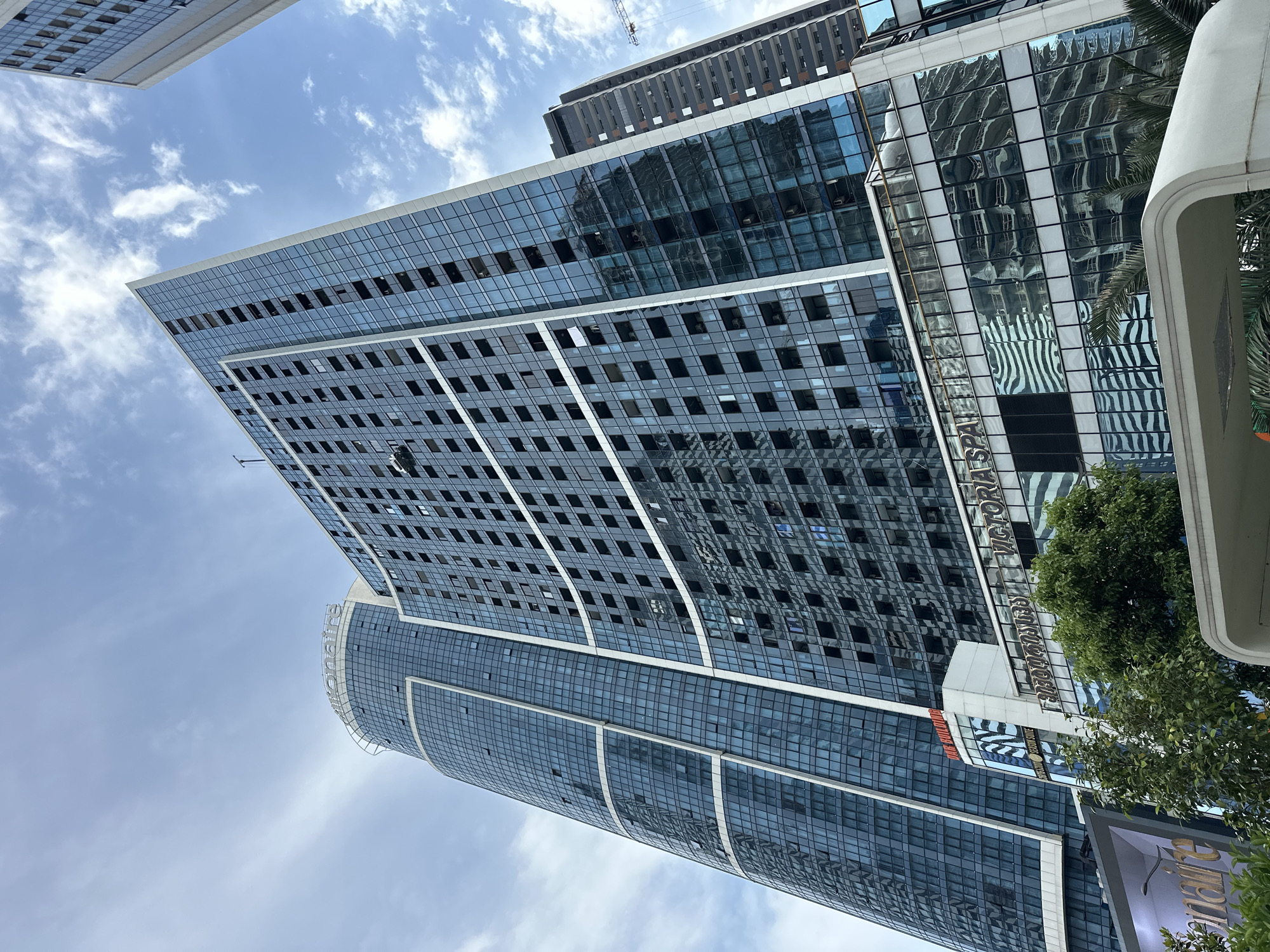 The image shows a modern skyscraper with a blue glass facade in Batumi, Georgia. The skyscraper is tall and angular, with many windows reflecting the sky and clouds. The text "Billionaire" is visible at the top of one of the buildings. Below the skyscraper, there are lower buildings with glass facades and the text "VICTORIA SPA" and "VICTORIA". Trees and palm trees are present in the foreground. The sky is partly cloudy.