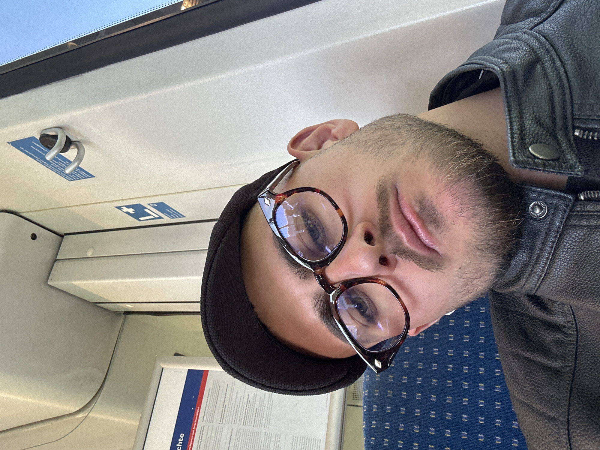 This image is a portrait-style shot of a man, likely a selfie, taken inside a train carriage. The original image is rotated 90 degrees clockwise; when viewed upright, the man's face fills a significant portion of the frame, looking directly towards the camera.
The subject is a man with short facial hair/stubble. He is wearing a black flat cap and a black leather jacket, visible on his right shoulder and arm. He has distinct tortoiseshell-framed glasses with visible reflections, suggesting light sources in front of him. His expression appears neutral and calm, with his eyes open.
The setting is the interior of a public transport vehicle, specifically a train, as indicated by the various details and the location context of Geldern, Germany. The background shows the train's internal structure: white/off-white ceiling panels on the left with a bright window area at the very top, suggesting daylight. A silver, double-pronged hook is visible on the ceiling panel, along with small blue stickers, one of which appears to have a cross symbol. On the right and below the man is blue patterned seat upholstery. A white information board or poster is partially visible below the man's chin.
Visible text on this information board includes "chte" (possibly part of a word like "Rechte" or "Geschichte") and "rsummis von Anschlußzügen," which translates to "delay of connecting trains." This text confirms the German context and indicates the poster likely pertains to travel information or passenger rights. The small blue stickers on the ceiling have illegible text but show graphic symbols.
The bright light from the top window suggests it is daytime. The scene captures a quiet moment of travel.