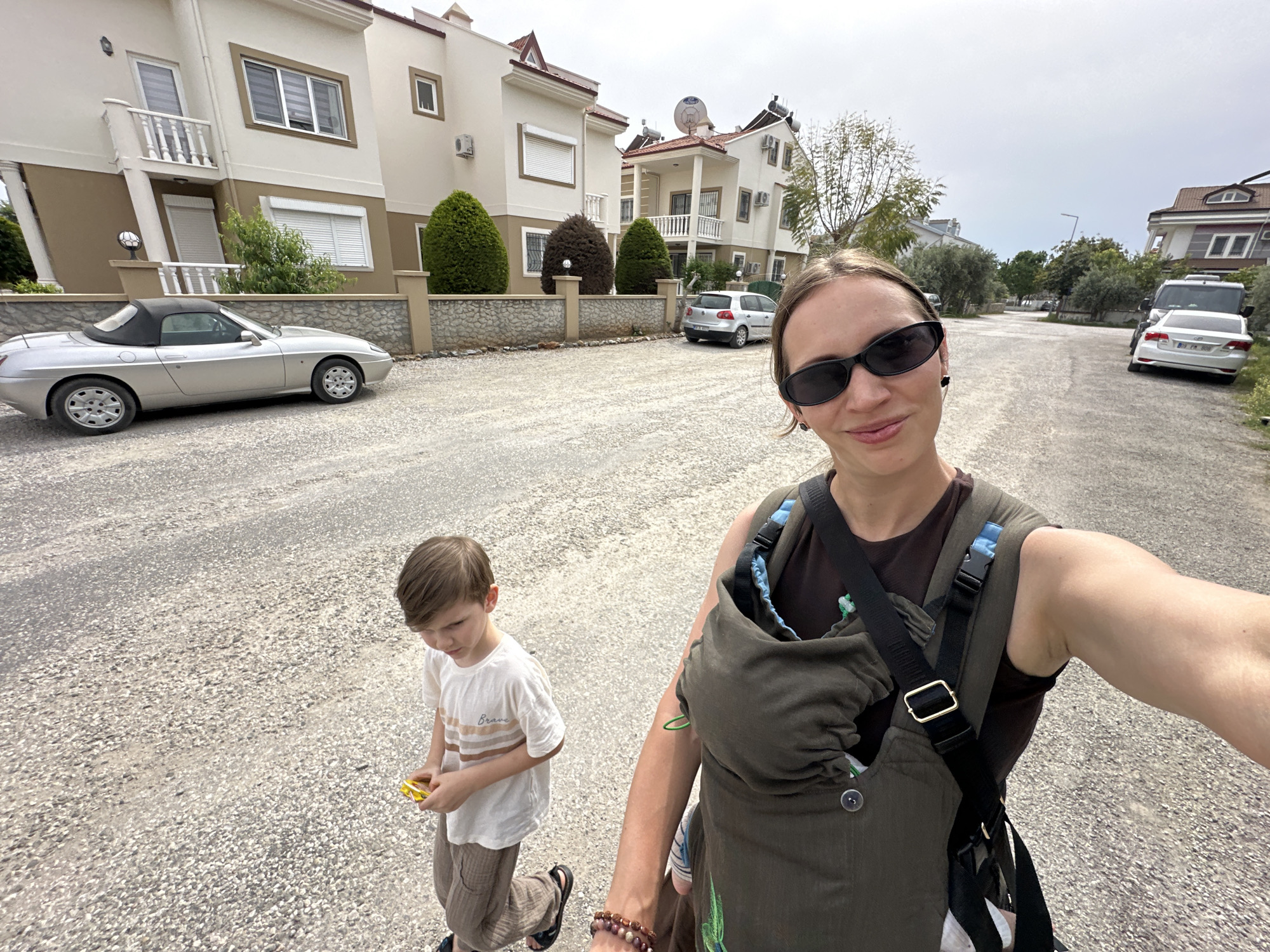 A street scene captures a woman holding a smartphone, posing for a selfie with a young boy walking beside her. The woman wears sunglasses and carries a baby in a green front carrier with black straps. The boy, dressed in a t-shirt and trousers, holds a yellow object.
The backdrop features houses with white and tan exteriors, set behind low stone walls. Several cars are parked along the street, including a silver convertible on the left. The street is paved with small stones. The sky is overcast.
The photograph was taken in the city of Fethiye, Turkey.