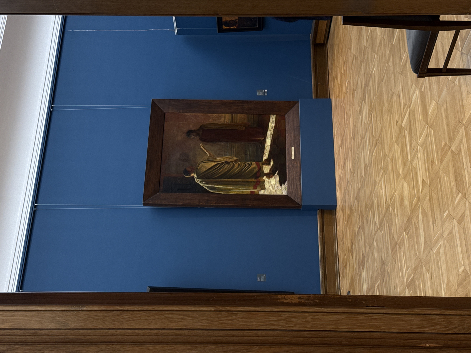 A framed painting of two figures in togas hangs on a dark blue wall in an art museum in Moscow, Russia. The wooden frame of the painting is darker than the oak wood trim along the bottom of the image. Above the painting are display wires hanging from the ceiling. A chair leg is visible in the upper right corner of the image. The museum has a wooden floor with a repeating diamond pattern.