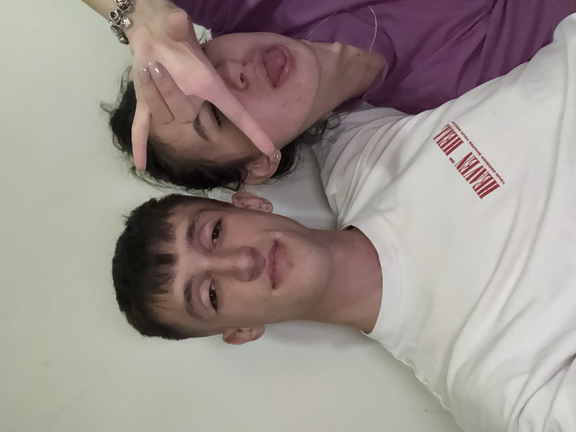 A close-up shot presents two individuals. The person in the foreground is wearing a white t-shirt with red text reading "HEAVEN HELL". The second person, positioned above and behind, is wearing a purple shirt, has dark hair, and is sticking their tongue out while making a "peace sign" with their hand obscuring part of their face. No background details are visible, and there are no indications suggesting a location within Stara Zagora, Bulgaria.