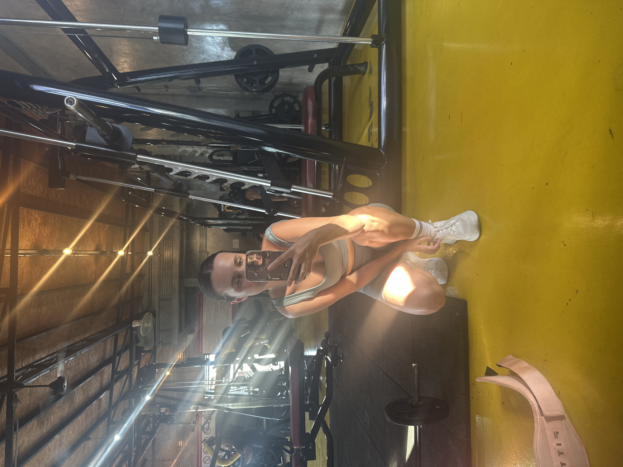 In the city of Baan Mae Nam, Thailand, a woman is captured in a mirror selfie inside a gym. She is in a squatting position, wearing a light green sports bra and matching shorts, white socks, and white athletic shoes. Her hair is pulled back into a bun, and she is holding a smartphone in her right hand, using it to take the photo. The gym environment is evident with various exercise equipment visible in the background, including weight machines, benches, and barbells. Sunlight streams in, creating dramatic light rays across the scene, illuminating the yellow-painted floor and the metallic structures of the gym equipment. A pink weightlifting belt is laid out on the floor in the foreground.