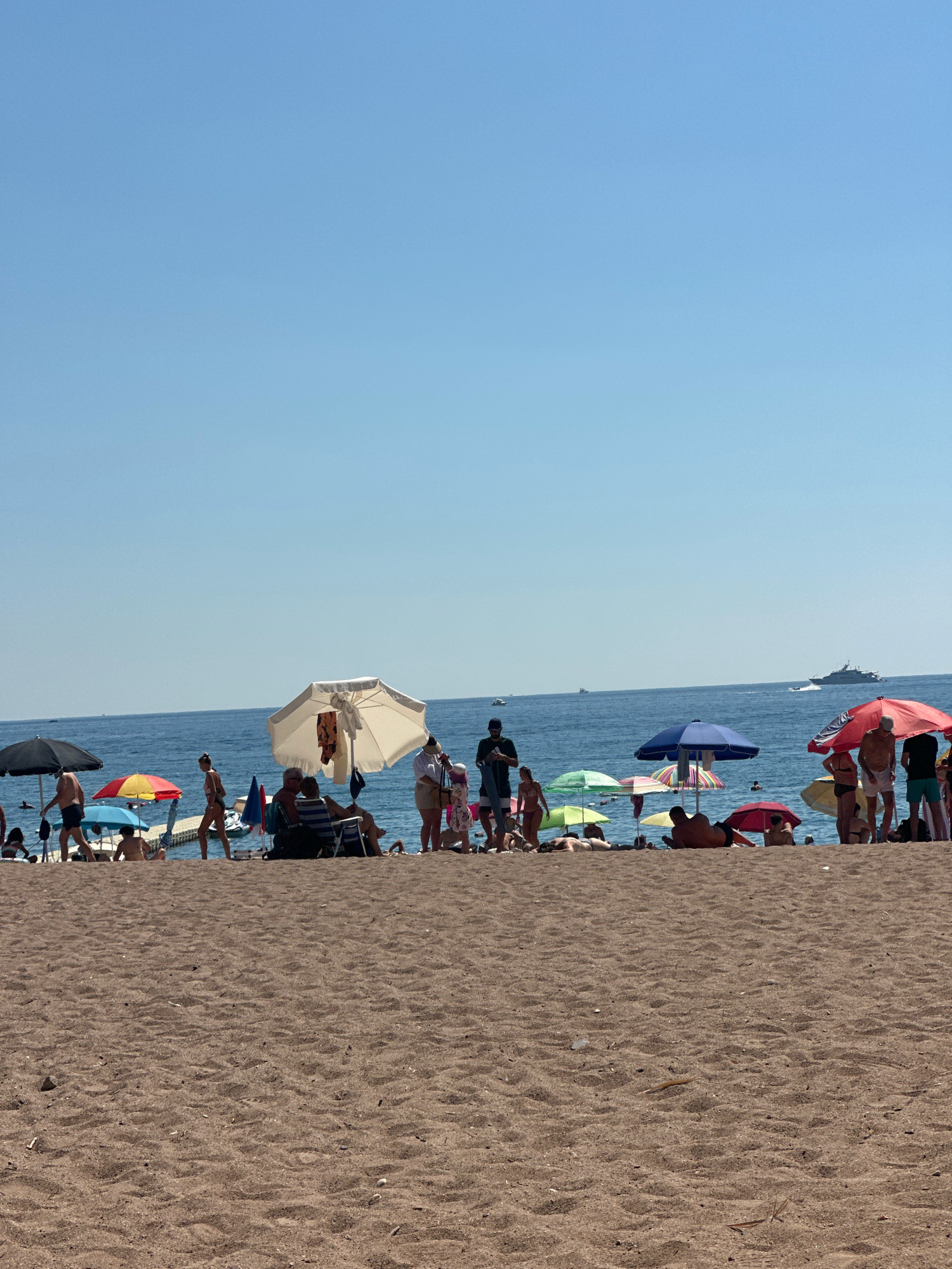 The image captures a crowded beach in Boreti, Montenegro, under a clear blue sky. A sandy foreground leads to the water's edge where numerous beachgoers are enjoying the sun and sea. Various colorful beach umbrellas, both open and closed, are dotted across the sand, providing shade for people lounging on chairs or towels. Several individuals are visible in the water and walking along the shoreline. In the background, the calm, blue sea stretches to the horizon, with a few boats visible in the distance. The overall scene depicts a typical summer day at a popular seaside resort.