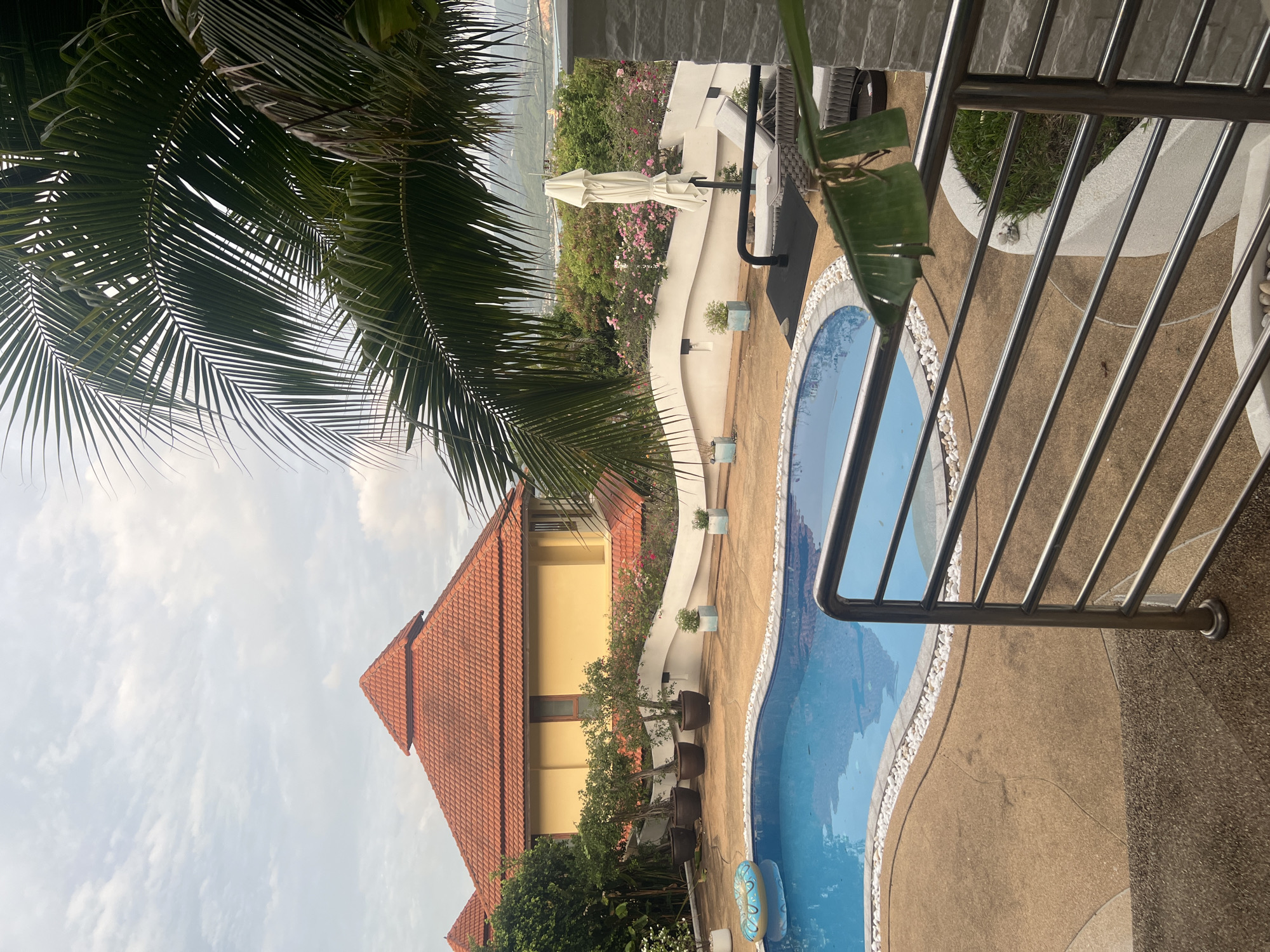 An outdoor swimming pool area at a resort or private residence in Ko Samui District, Thailand, features a kidney-shaped pool with blue water and a light blue inflatable ring. The pool is surrounded by a tan-colored concrete deck with white pebble accents along its edge. To the left of the pool, a building with a distinctive orange-tiled, multi-tiered roof and yellow walls is visible, partially obscured by lush green foliage. Several potted plants, including small trees in dark brown planters, are arranged along a curved, off-white wall that separates the pool area from a garden. Beyond this wall, a variety of flowering plants, predominantly pink and white, are in bloom. A closed white patio umbrella stands next to a lounge chair with gray cushions. In the foreground, a silver metal railing and concrete steps lead down towards the pool area. Palm fronds hang into the frame from the top right, casting shadows. The sky is overcast, with a mix of clouds and lighter patches. In the distance, rolling green hills and glimpses of what appear to be other buildings and water bodies can be seen. The overall scene depicts a tropical setting, likely during the day.