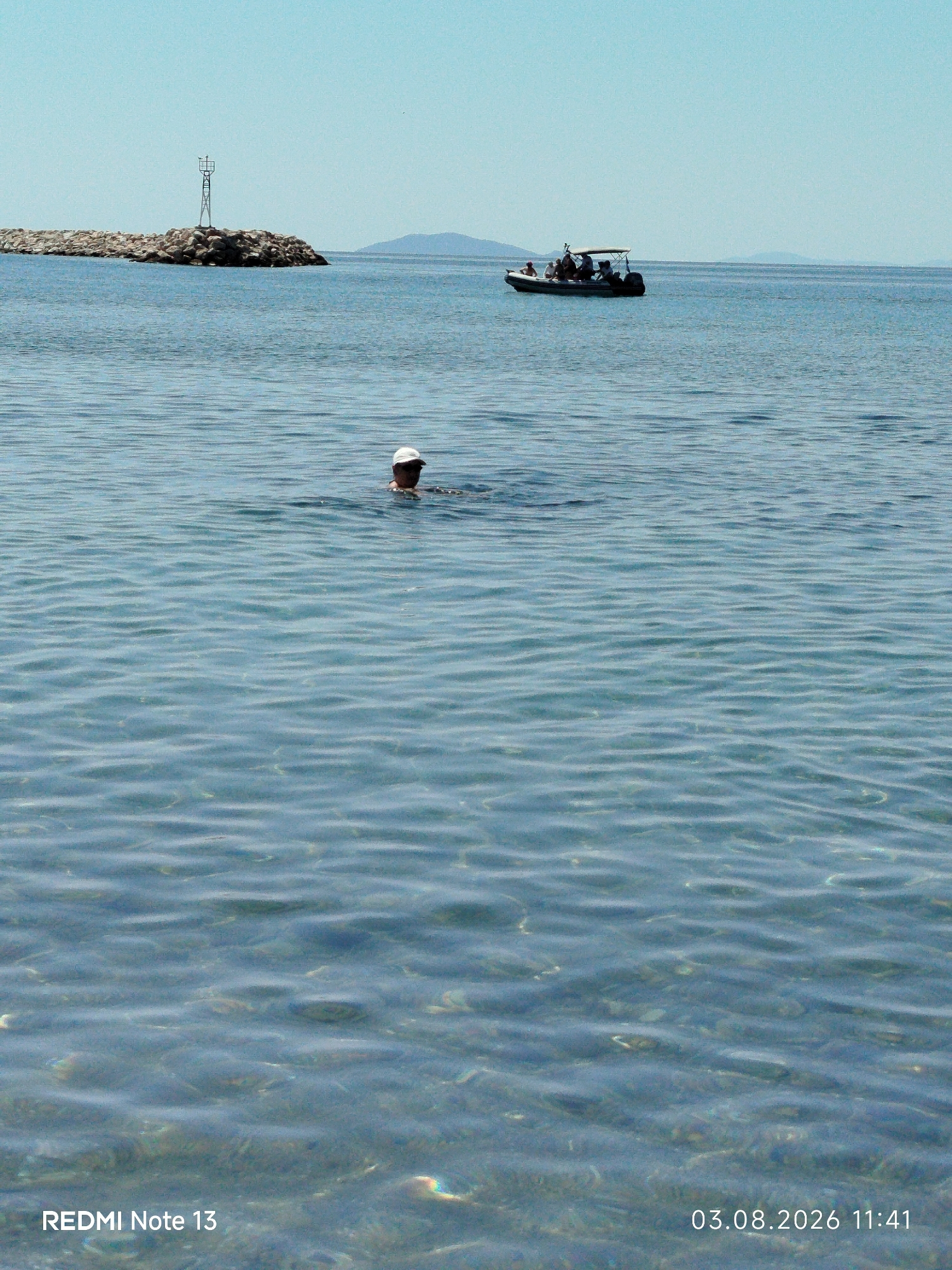 In the clear blue waters of Sithonia Municipal Unit, Greece, a solitary individual wearing a white cap swims. Their head and shoulders are visible above the gently rippling surface. In the distance, a small boat carrying several passengers navigates the sea. To the left, a man-made breakwater, constructed from large stones, extends into the water, topped with a navigational marker. The horizon is marked by distant, hazy hills under a pale blue sky. The seabed is visible through the transparent water, revealing pebbles and the subtle play of light on the sandy bottom. The timestamp "03.08.2026 11:41" and the text "REDMI Note 13" are overlaid on the lower portion of the image.