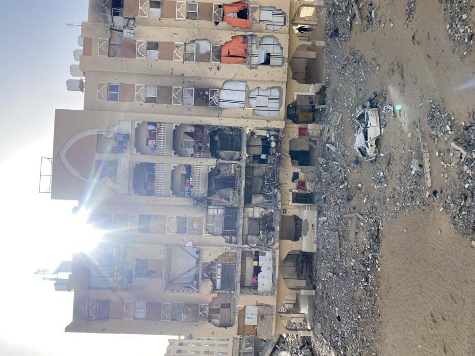 The image displays a multi-story building in Khan Yunis, Palestinian Territory, exhibiting extensive structural damage under bright sunlight. The light-colored facade is heavily pockmarked with numerous small impacts and has multiple large sections blown out, exposing the interiors of various apartments across several floors. Remnants of furniture, draped fabrics, and articles of clothing are visible within these exposed sections and on damaged balconies. Charring is evident on the right side of the building's upper floors. The rooftop features visible antennas and water tanks. The ground level in front of the building is covered with a thick layer of concrete rubble, sand, and twisted metal debris. In the mid-ground, a severely damaged, white four-door sedan, crushed and missing its roof and other major components, rests amidst the rubble. A single individual, dressed in dark clothing, is observed walking from right to left along the ground level near the base of the building on the right side.