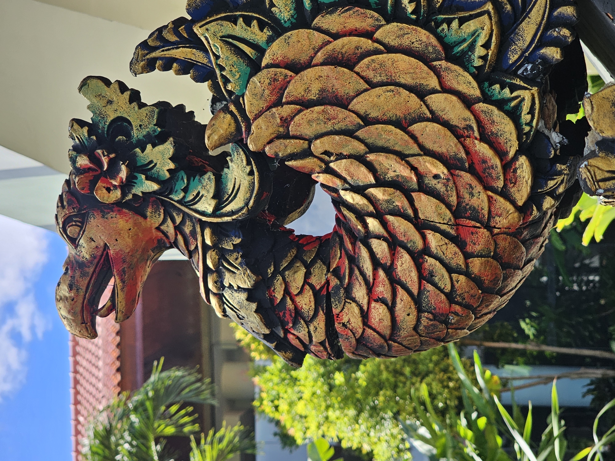 The image depicts a brightly colored, stylized Garuda statue. The statue has a bird-like head with an open beak, a body covered in scale-like patterns colored in gold and red, and decorative foliage elements in blue and green. The statue is situated outdoors, possibly attached to a building, as evidenced by the partial view of a white wall and eaves in the background. The background also contains greenery, including trees and smaller plants. In the distance, part of a building with a red-tiled roof is visible against a blue sky. The location is Kuta Selatan, Indonesia.