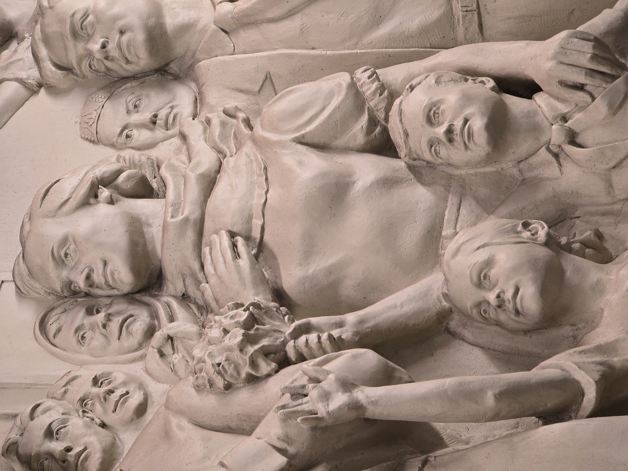 This image is a close-up of a bas-relief sculpture in Unknown, Unknown. The sculpture depicts a group of human figures, seemingly lying down or collapsed together. The figures, rendered in a pale, stone-like material, have detailed facial features and appear to be in various states of rest or repose. One figure, situated towards the center of the image, seems to be embracing or holding a bouquet of flowers. The surrounding figures are positioned closely, some with arms intertwined, creating a sense of unity and potentially sorrow or collective grief. The overall texture is rough and unfinished, adding to the solemn and somber tone of the sculpture.