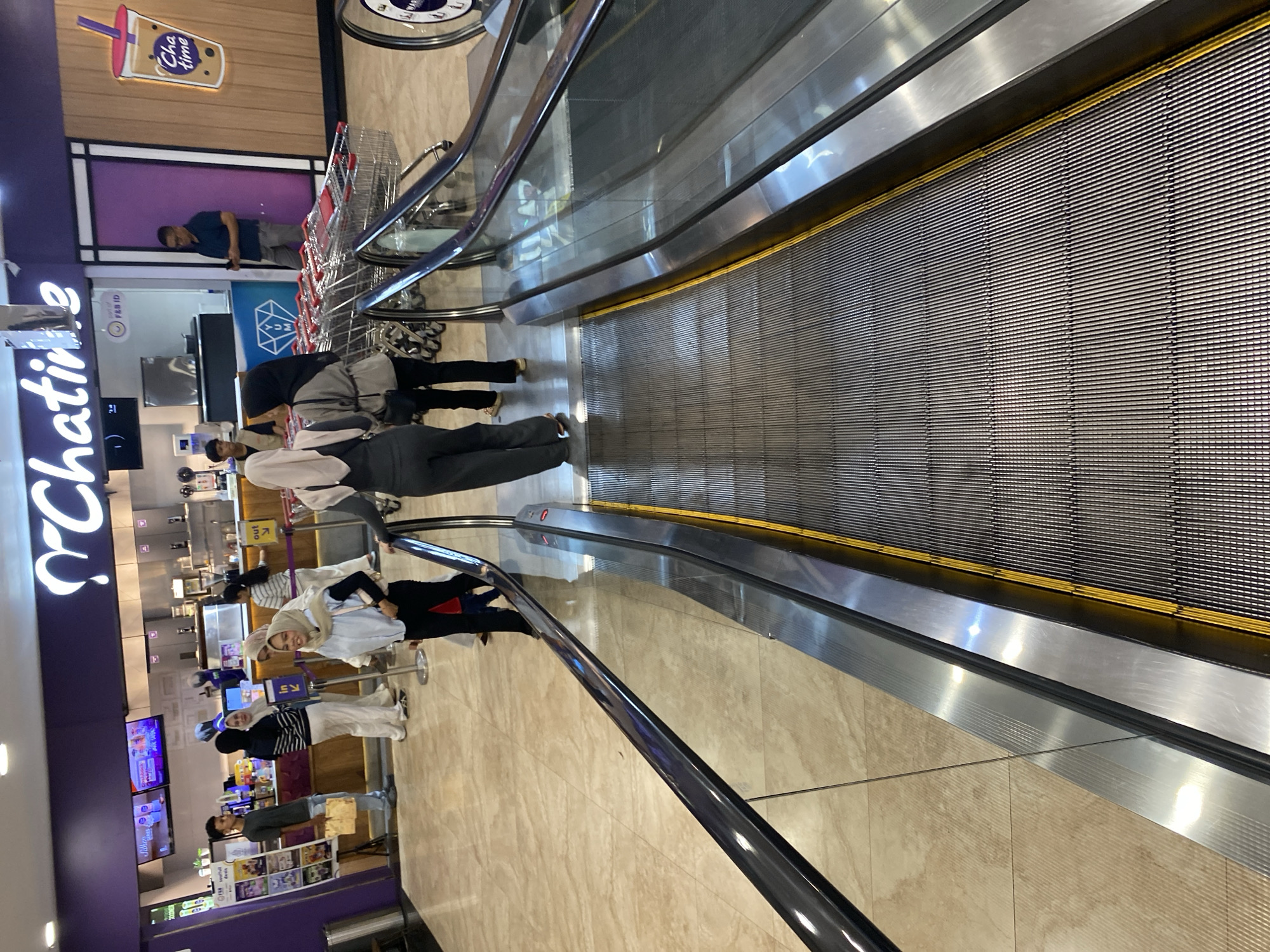 An exterior shot of a Chatime store in Banda Aceh, Indonesia, is captured from a slightly elevated perspective, facing an escalator. The store's purple and white signage is prominent above the counter area. A group of individuals, some wearing headscarves, are seen interacting and queuing at the counter. In the foreground, a man and a woman stand on the escalator, their backs to the camera, descending. Several shopping carts are neatly arranged to the right of the escalator. The floor is tiled in a light beige and cream pattern. The overall scene depicts a busy commercial environment.