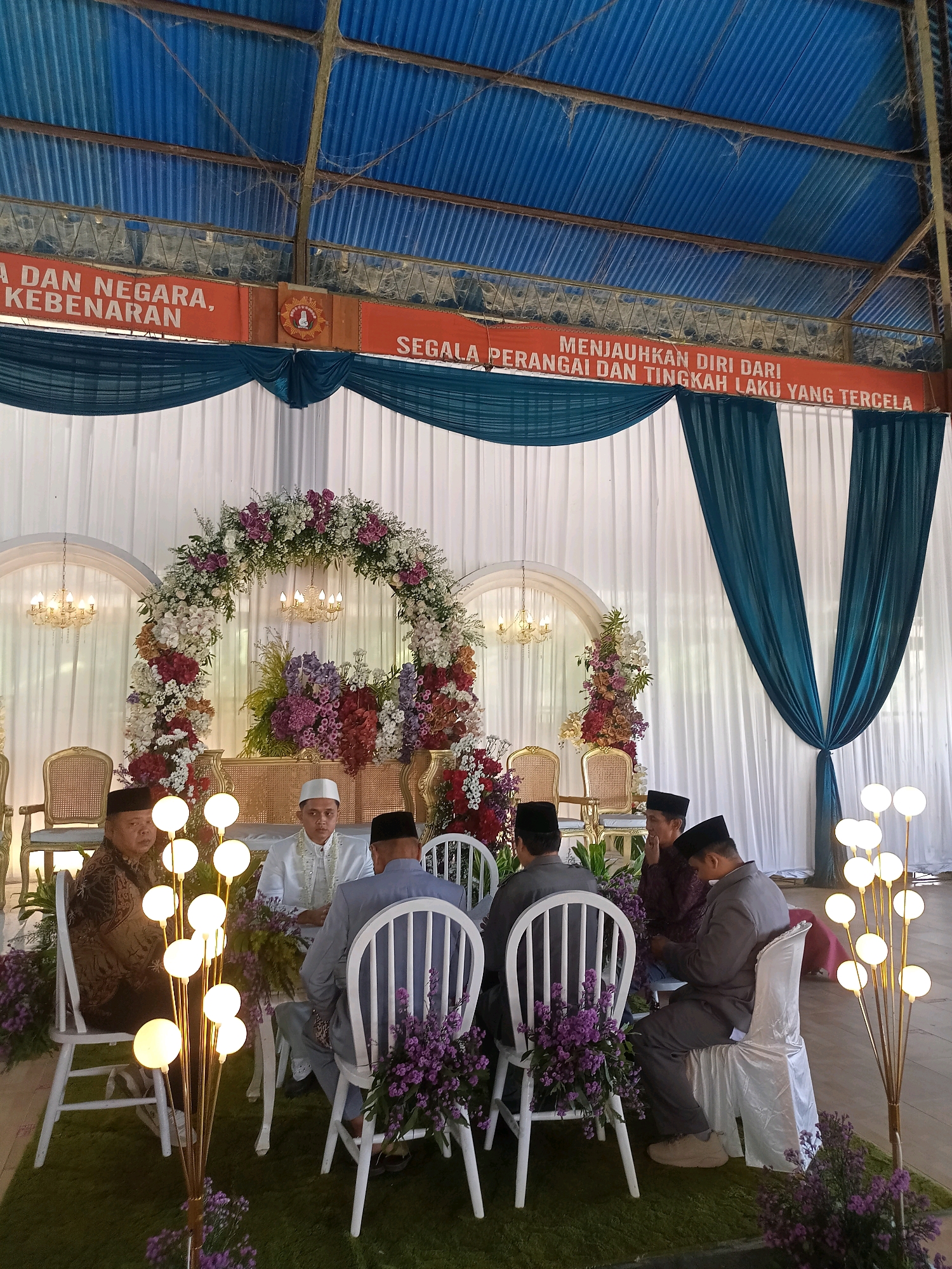 The image depicts an indoor event, likely a wedding reception, in Wanadadi, Indonesia. The venue appears to be a large hall with a corrugated blue roof. A banner with orange text, partially obscured by the roof structure, reads "A DAN NEGARA, KEBENARAN" on the left and "SEGALA PERANGHAI DAN TINGKAH LAKU YANG TERCELA" on the right. The backdrop features white draped fabric with teal fabric accents.
In the foreground, several men are seated on white chairs around tables. One man in the center, wearing a white cap and white attire, appears to be the central figure, possibly the groom. He is looking forward. To his left, a man in a patterned brown and black garment is seated, looking towards the center. To the right of the central figure, two men are seated, one in a gray suit looking down at his phone, and another in a darker garment looking towards the center. The chairs are decorated with white satin sashes and purple flowers.
A prominent floral archway, adorned with a variety of pink, white, and purple flowers, stands behind the seated individuals. Two ornate chandeliers hang from the ceiling within arched alcoves. The floor is covered in green carpet, and decorative floor lamps with multiple white globe lights are positioned throughout the scene. Purple flowers are also placed on the floor around the lamps. The overall impression is of a formal and decorated celebration.